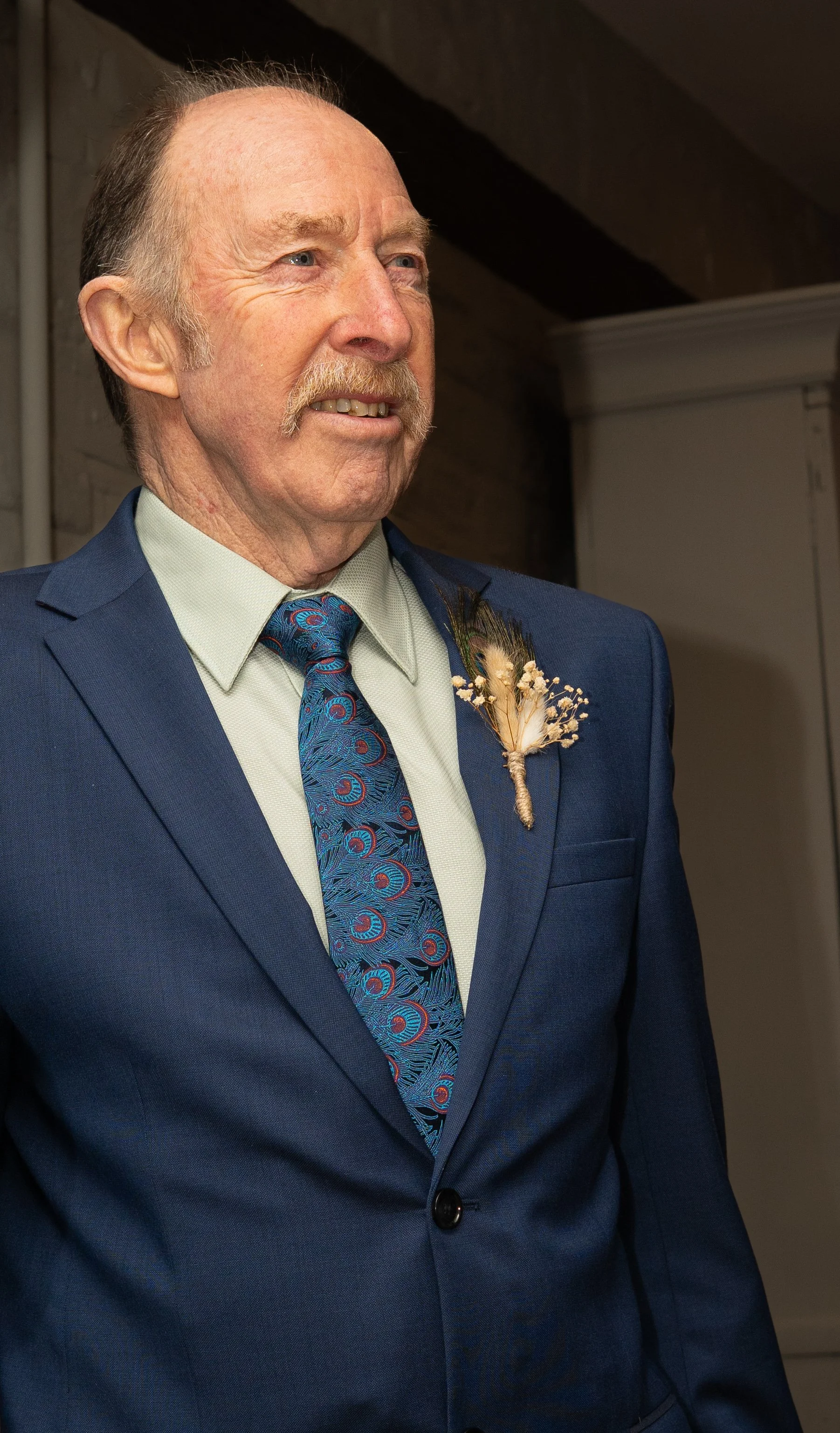 Close-up of an elderly man in a blue suit with a patterned tie, smiling, with a boutonnière pinned to his lapel.