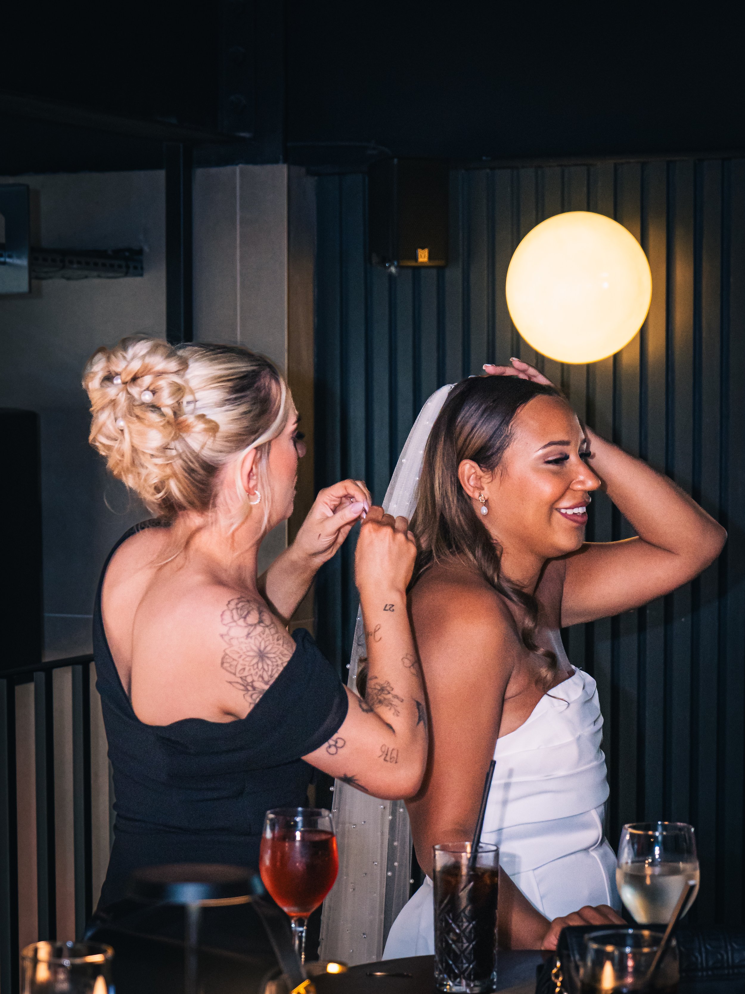 A woman in a white wedding dress is getting ready for her wedding, assisted by another woman with tattoos, in a dimly lit room with glasses of drinks on the table.