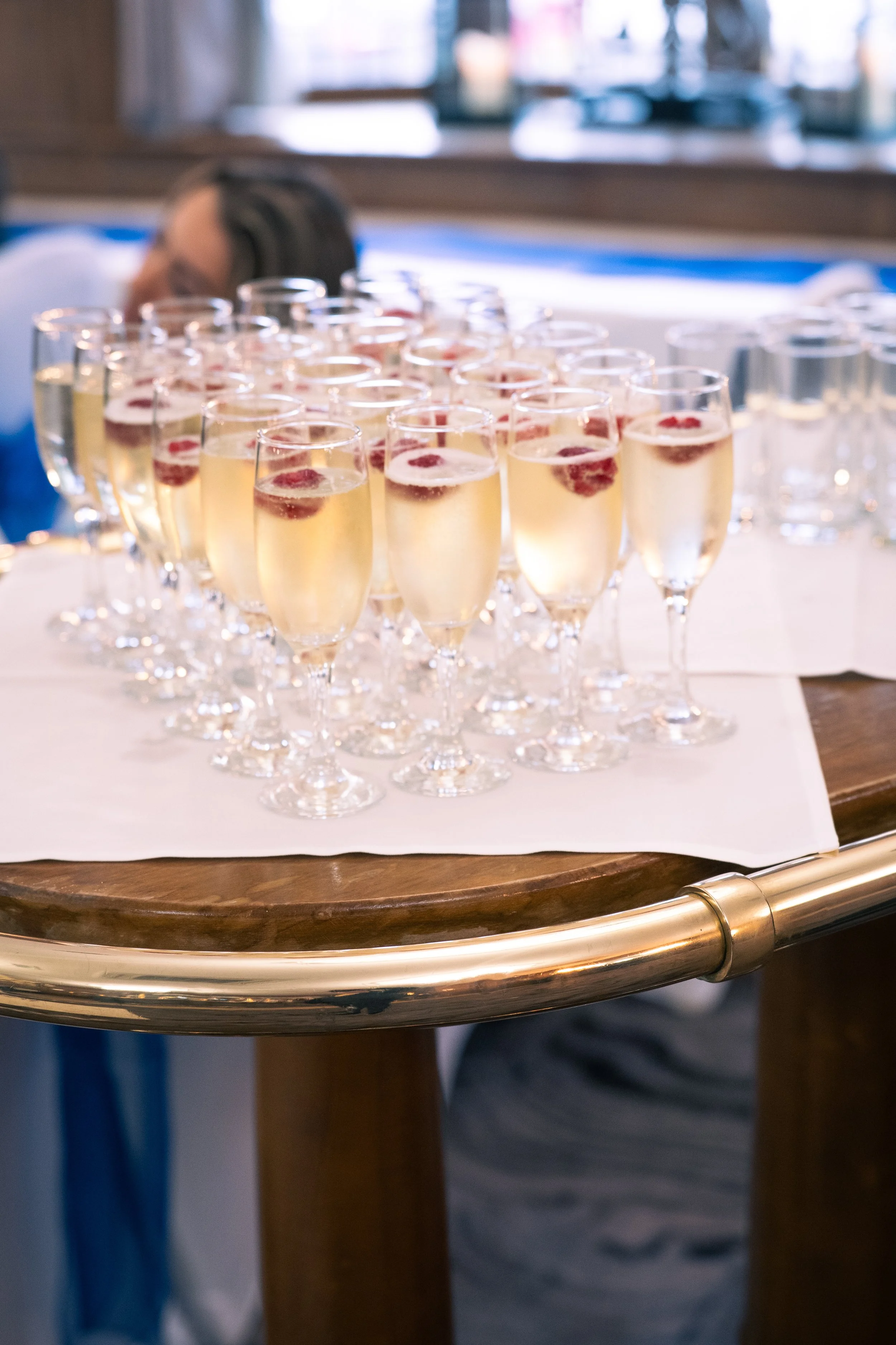 Multiple glasses of champagne with raspberries on top, arranged on a table with a white cloth.