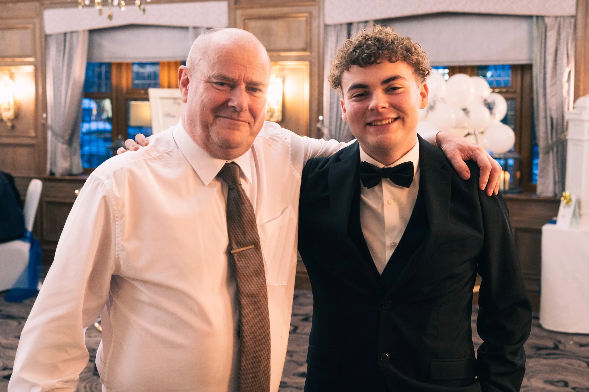 Two men dressed up at a formal event, standing together in a decorated room, smiling at the camera. The man on the left wears a white shirt and tie, the man on the right wears a tuxedo with a bow tie.