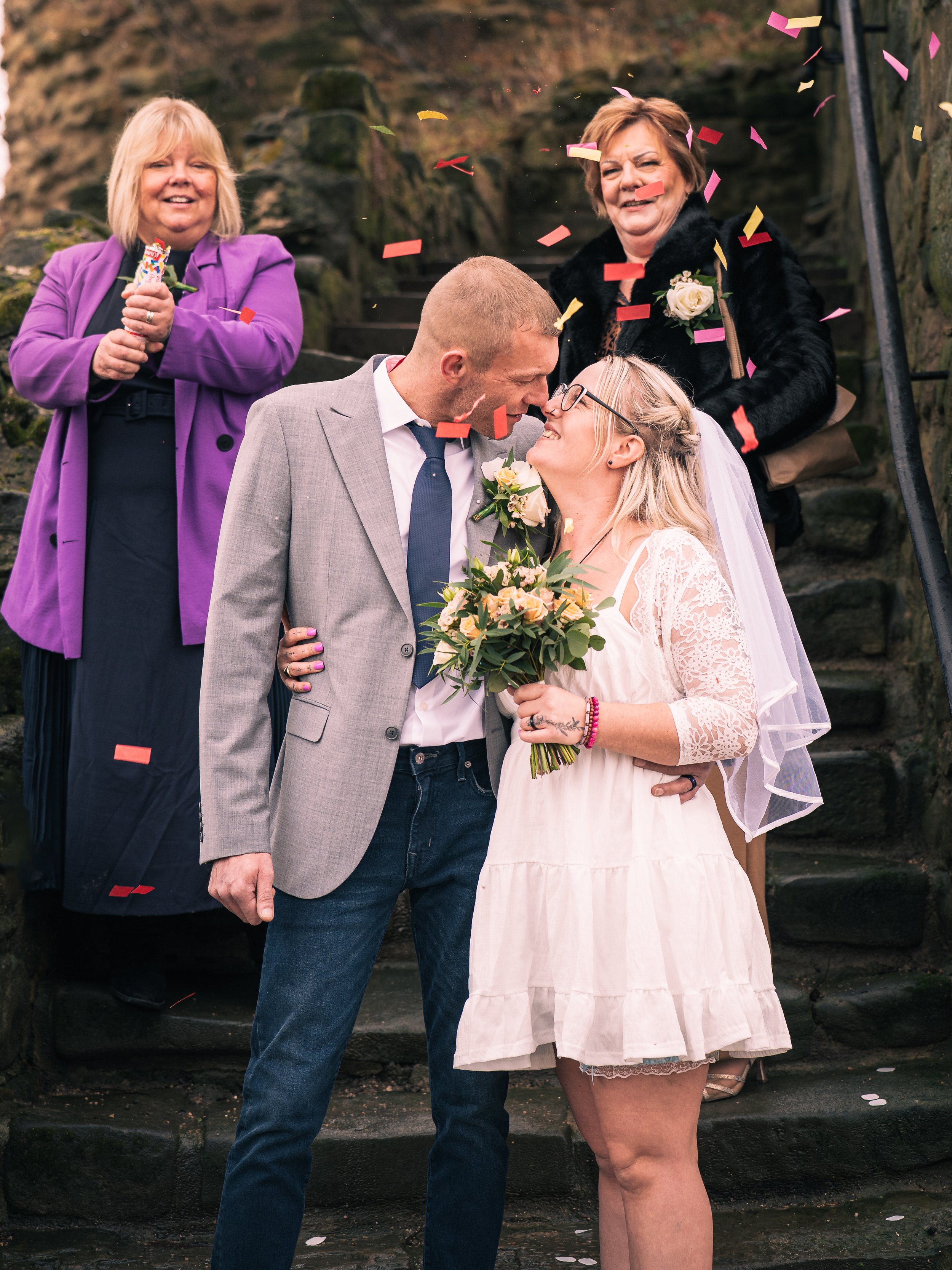 A couple getting married outdoors, leaning faces touching, surrounded by family and friends. Confetti falls around them as they celebrate their wedding on stone steps.
