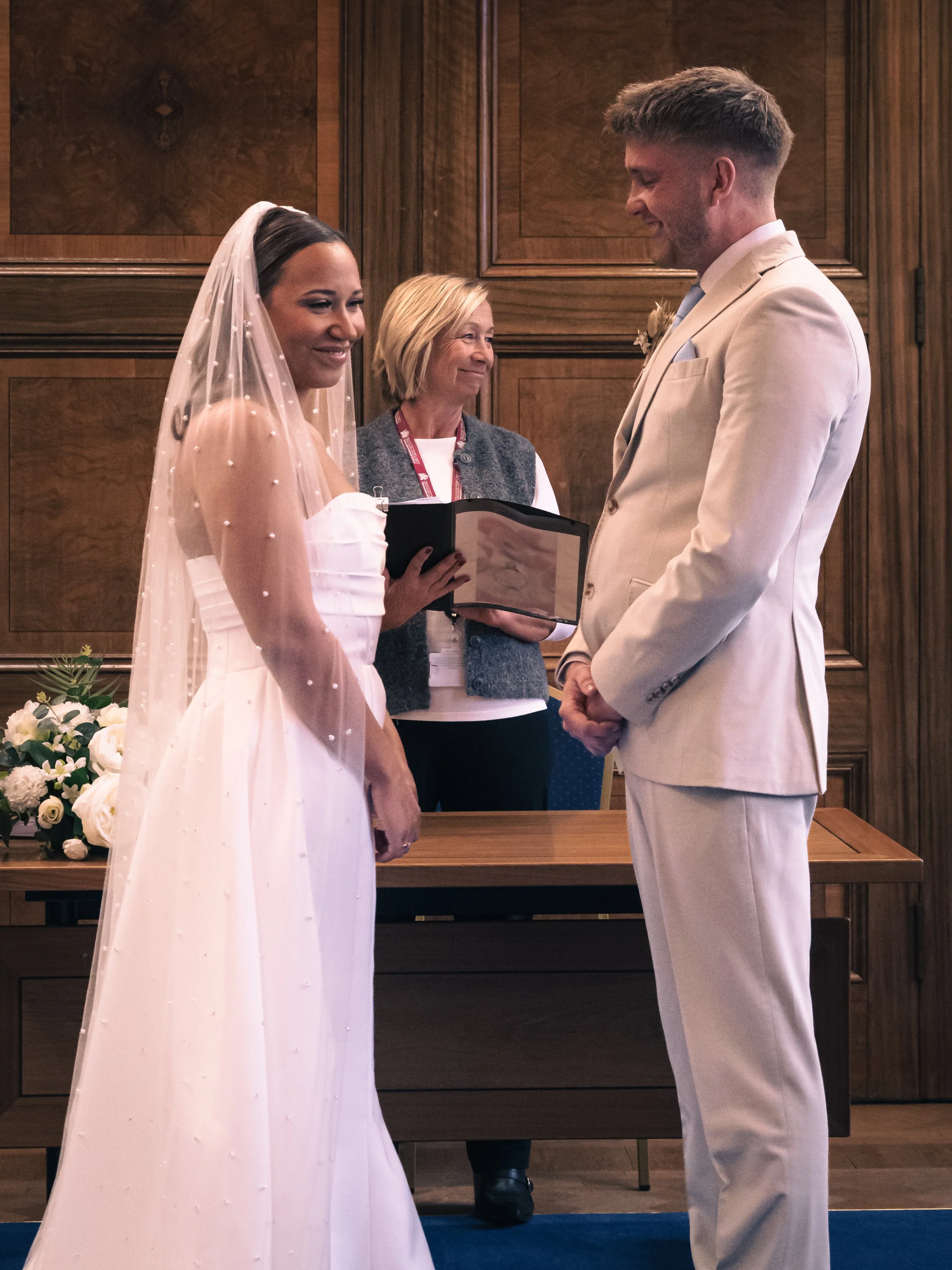 A bride and groom exchanging vows during a wedding ceremony, with a woman officiant standing behind them and floral arrangements on a wooden table in a wood-paneled room.