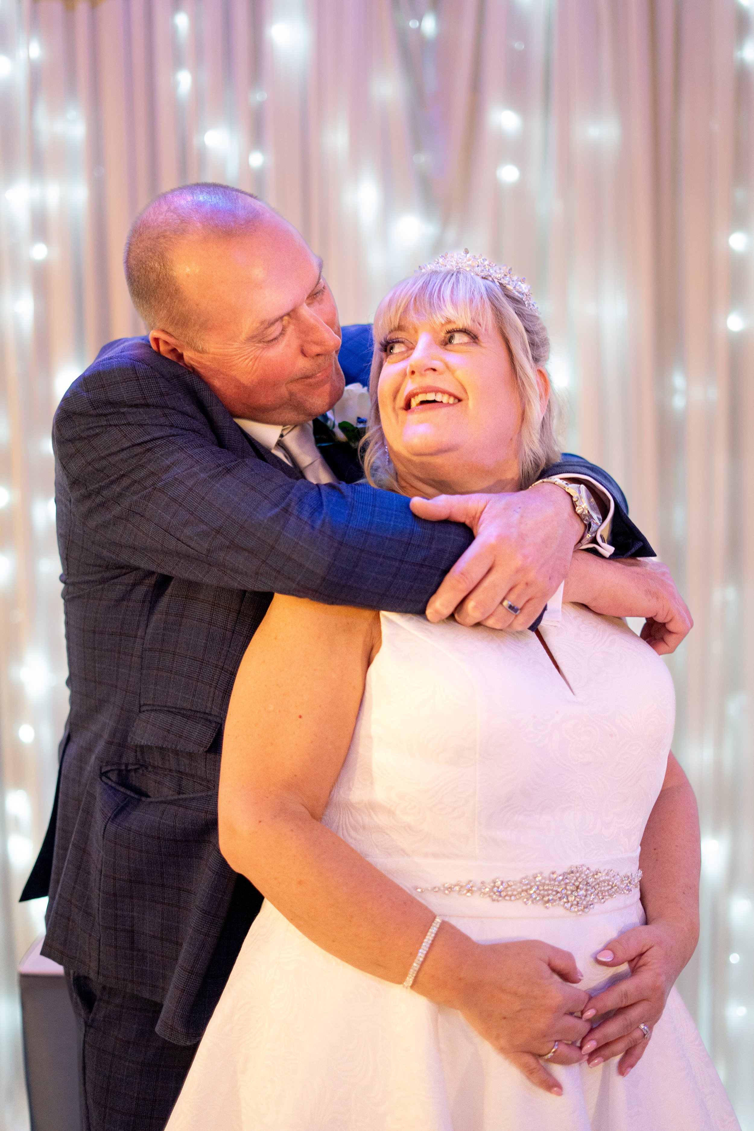 A happily married couple, a man in a suit and a woman in a wedding dress, sharing a joyful embrace at their wedding reception.