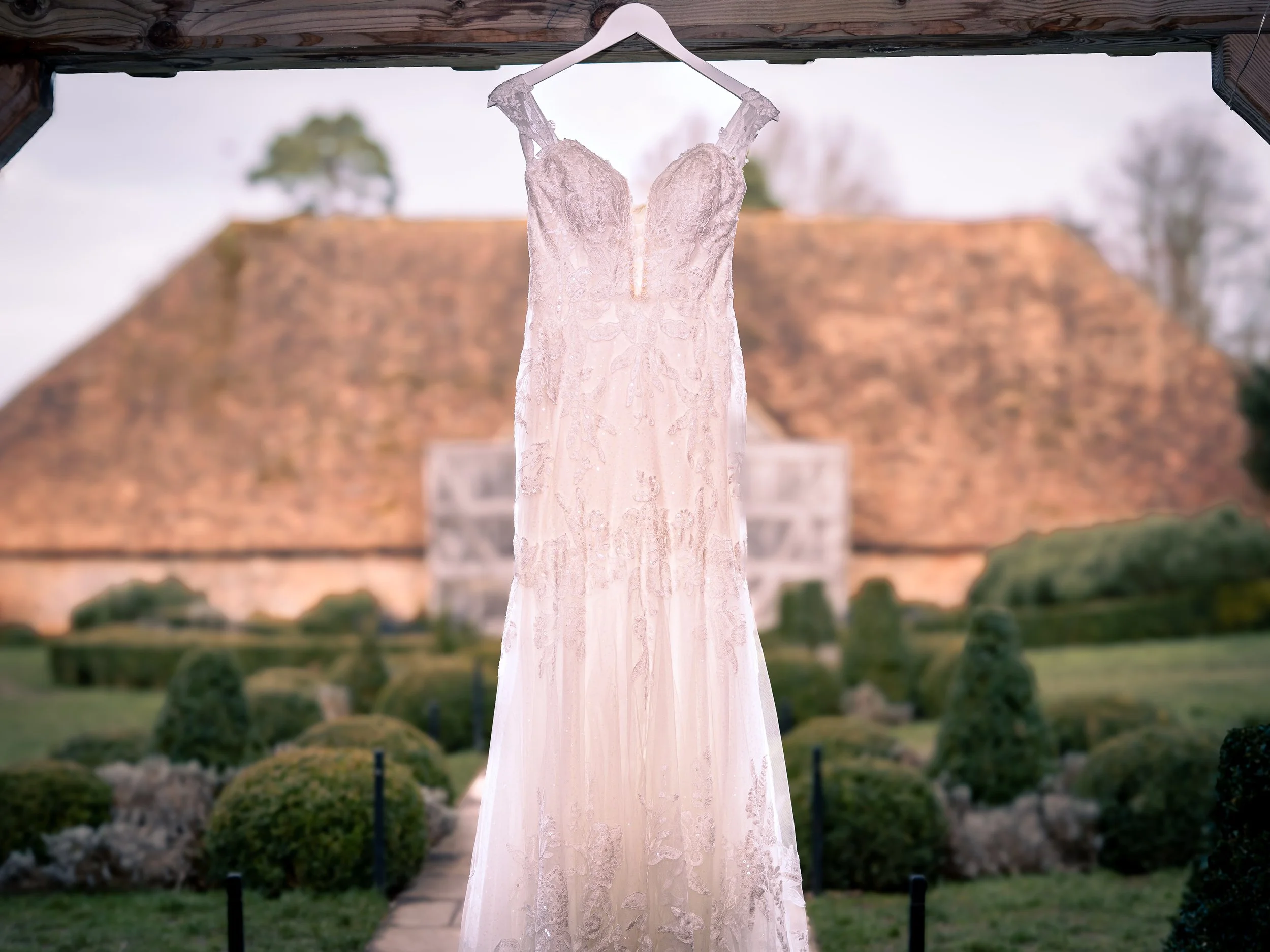 A wedding dress hanging from a hanger outdoors with a garden and a large brick building in the background.