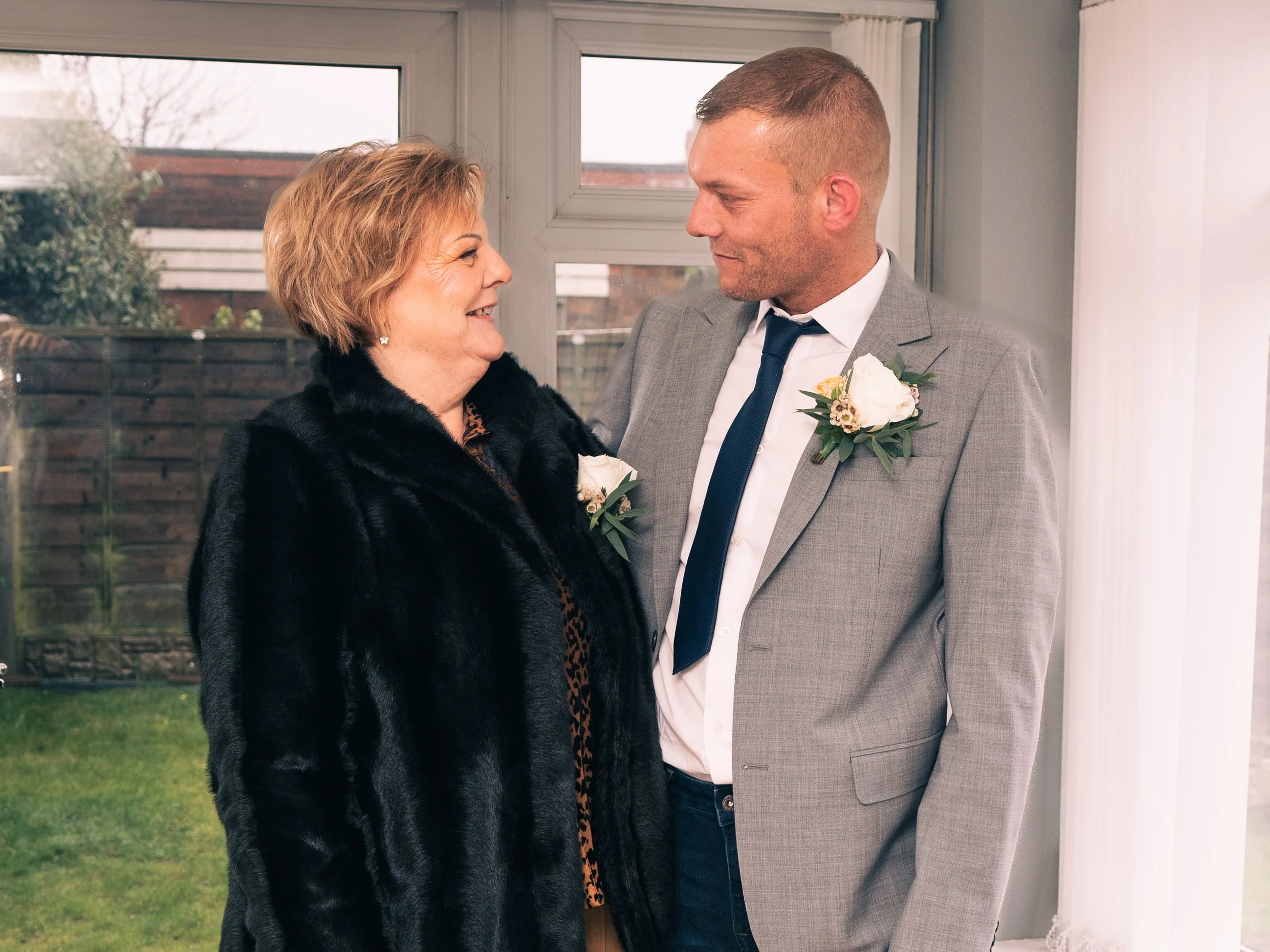 A woman and a man stand indoors, smiling at each other, dressed formally with boutonnières, likely celebrating a special occasion.