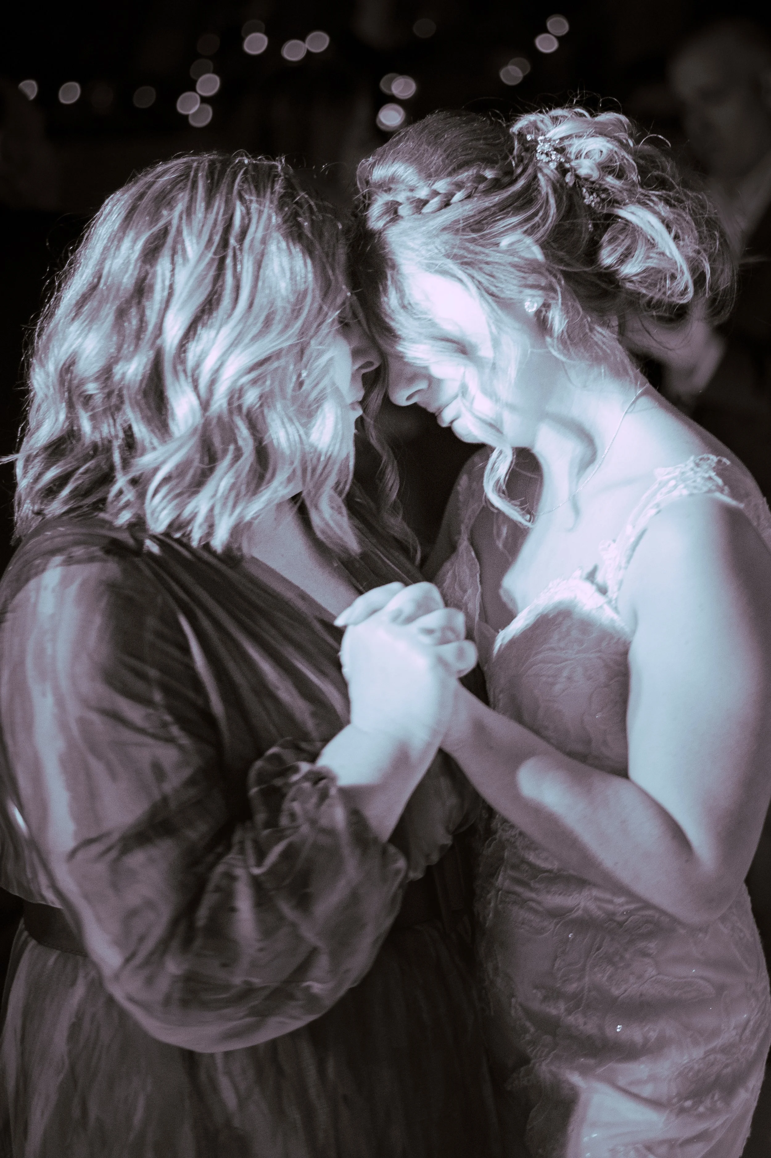 Two women sharing a dance, their foreheads touching and eyes closed, in a close embrace at a celebration or wedding, with blurred lights in the background.
