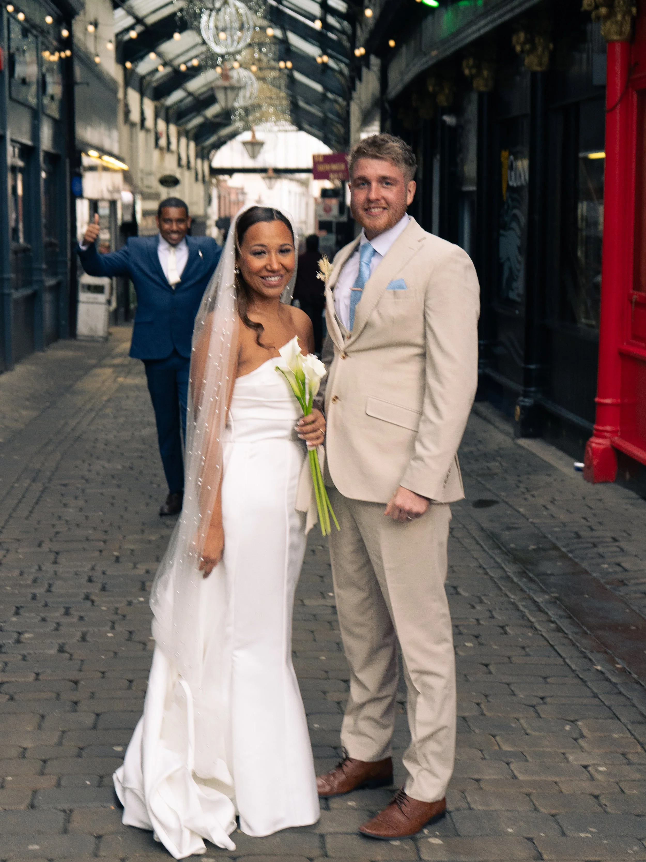 A bride and groom smiling at the camera while standing under a canopy on a cobblestone street. The bride is in a white strapless wedding dress, holding a small bouquet of white calla lilies, and wearing a veil. The groom is in a light beige suit with