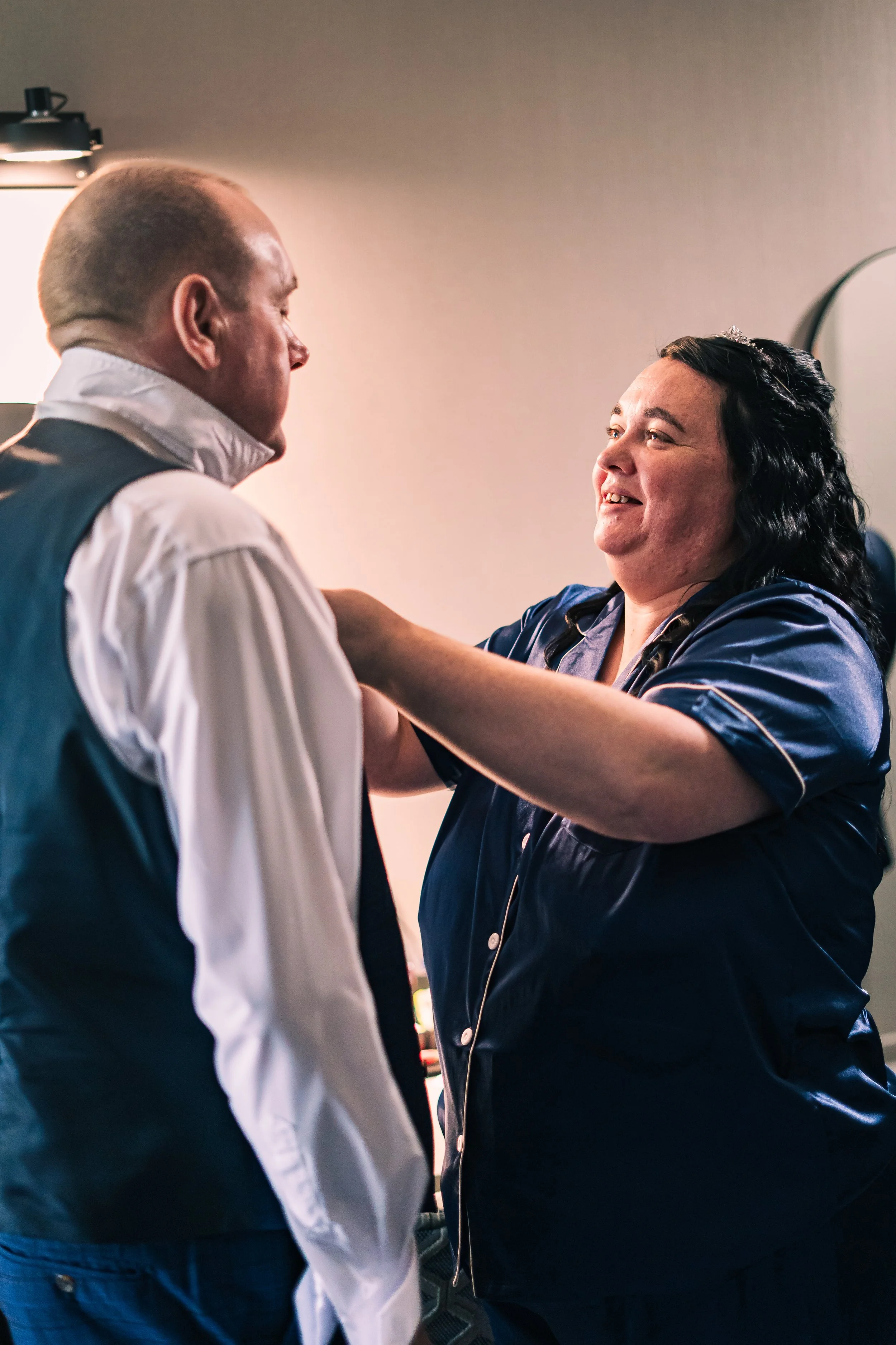 A woman in navy pajamas smiling as she helps a man in a white shirt and dark vest prepare for a wedding.