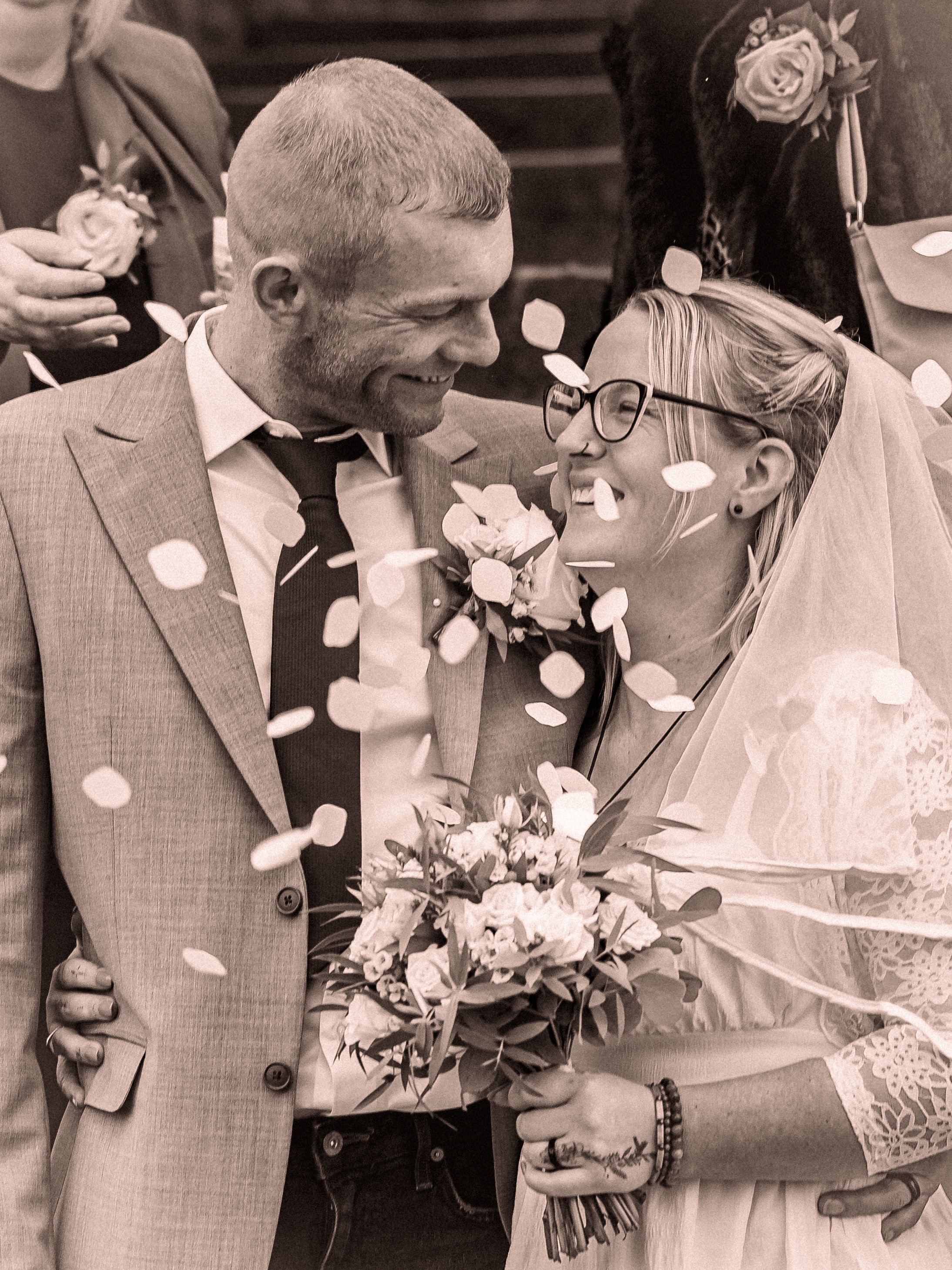 A couple sharing a joyful moment at their wedding, with the bride holding a bouquet and both smiling at each other amidst falling confetti.