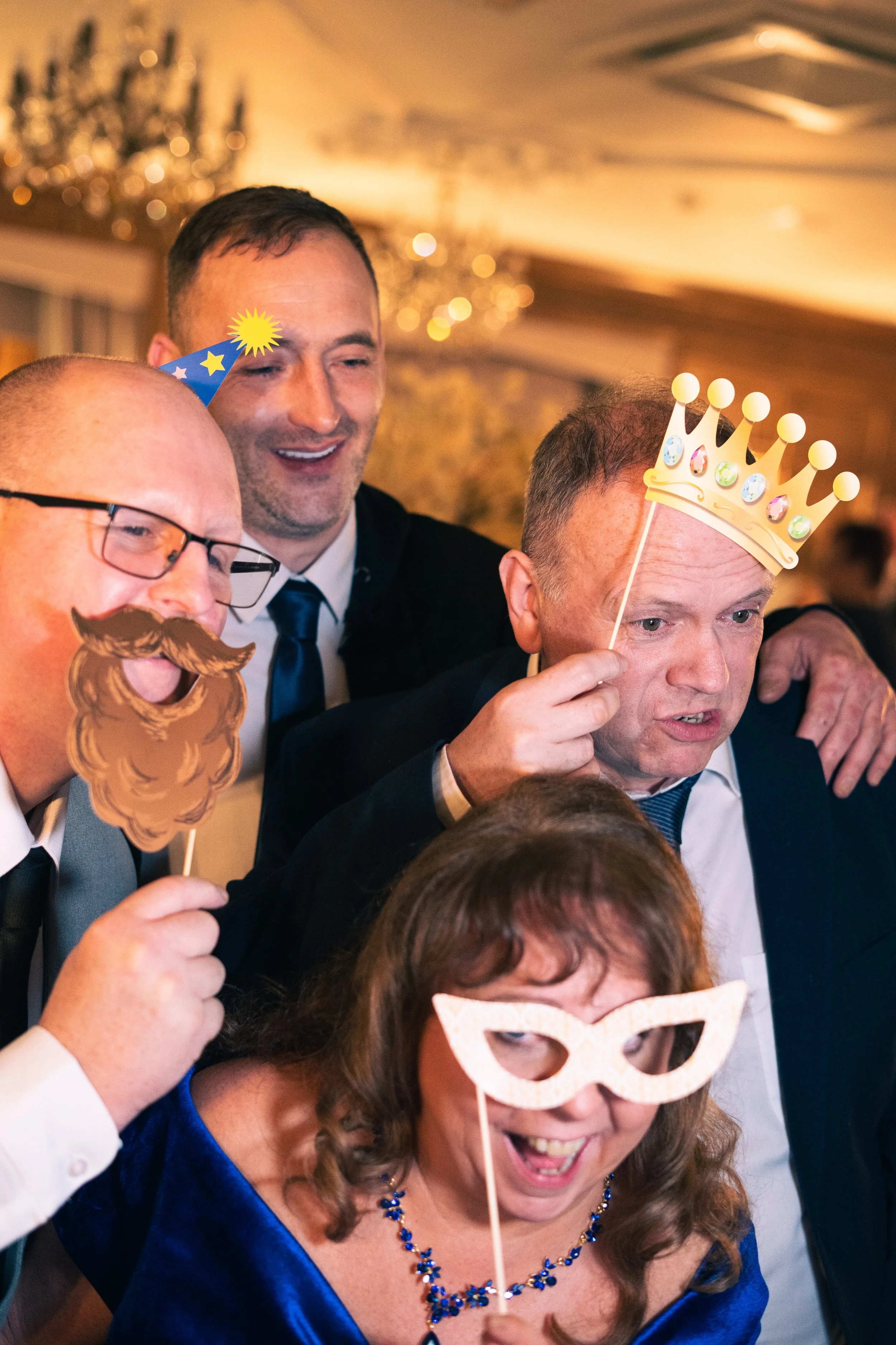 People at a celebration wearing party props: a man with a paper crown, another with a wizard beard, and a woman with a fancy mask.