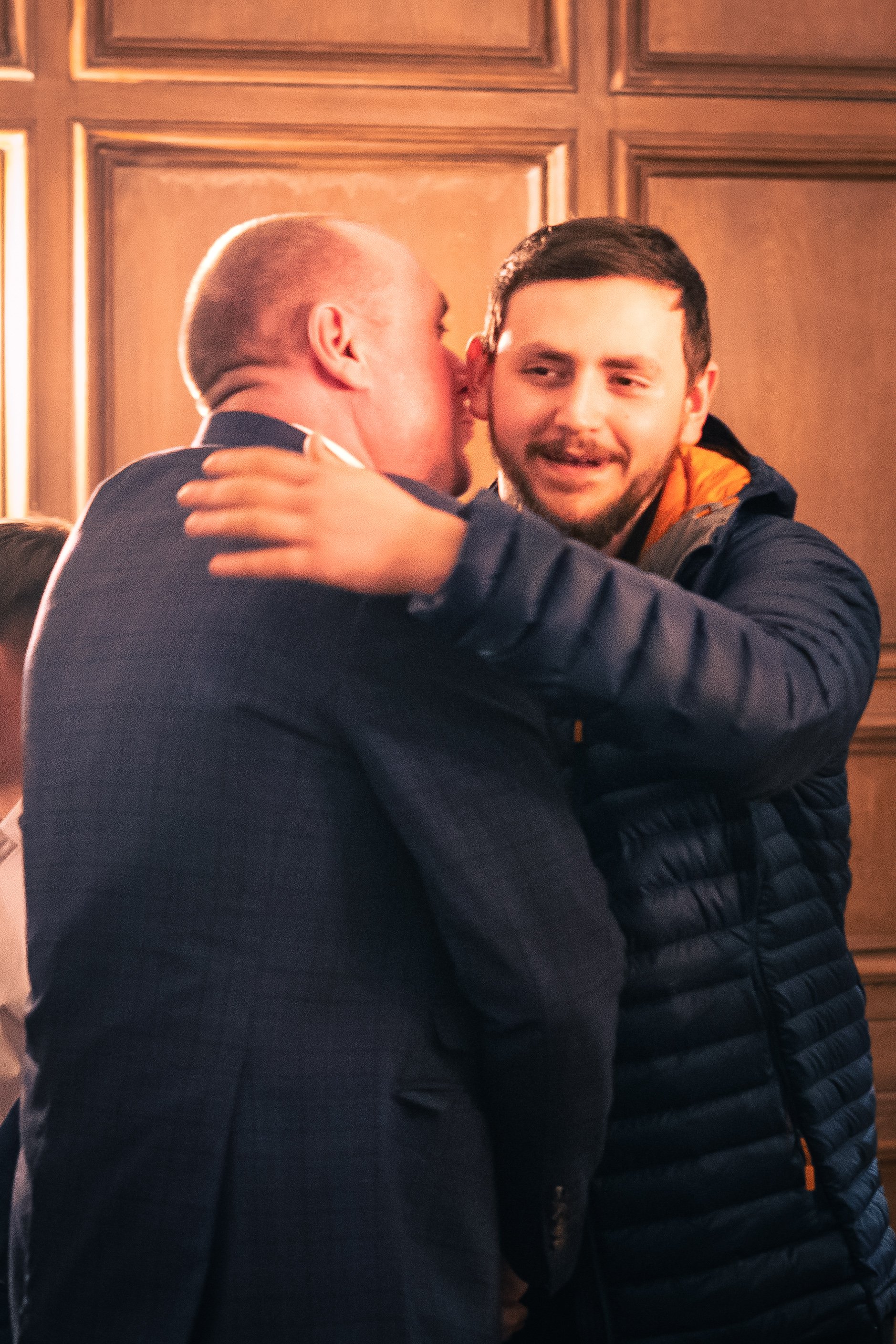 Two men embrace in a warm hug, one with his face close to the other's cheek, and the other smiling with closed eyes, in a room with wooden wall paneling.