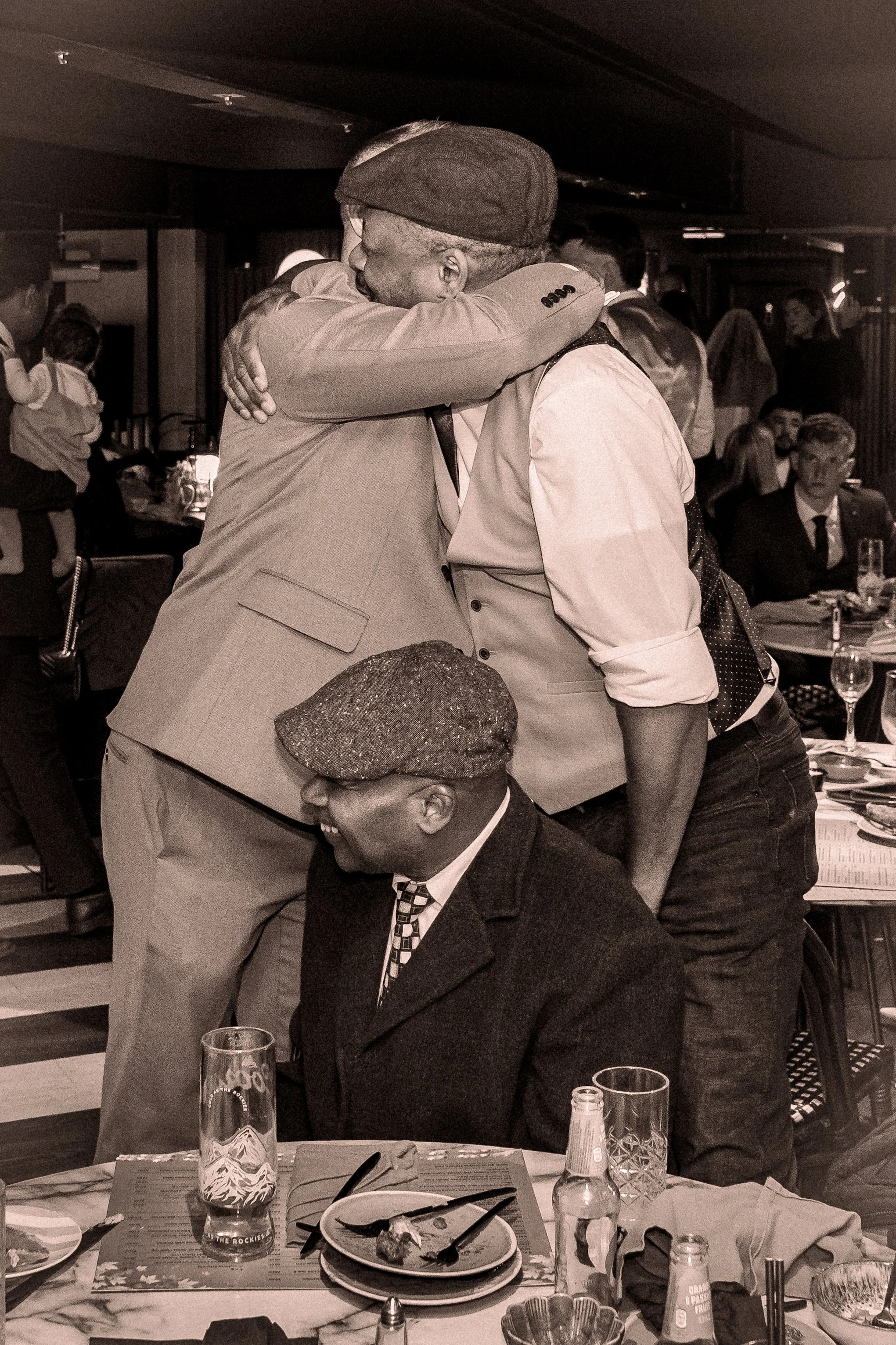 People hugging at a celebration or gathering in a restaurant or event space, with others seated and standing around in the background.
