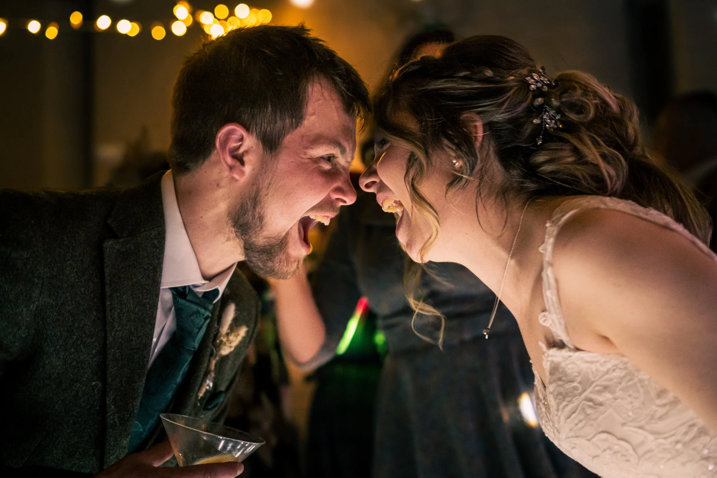 A man and a woman, both smiling and leaning their foreheads together at a celebration or party, with the woman wearing a white dress and the man dressed in a dark suit and tie. They appear happy and joyful.