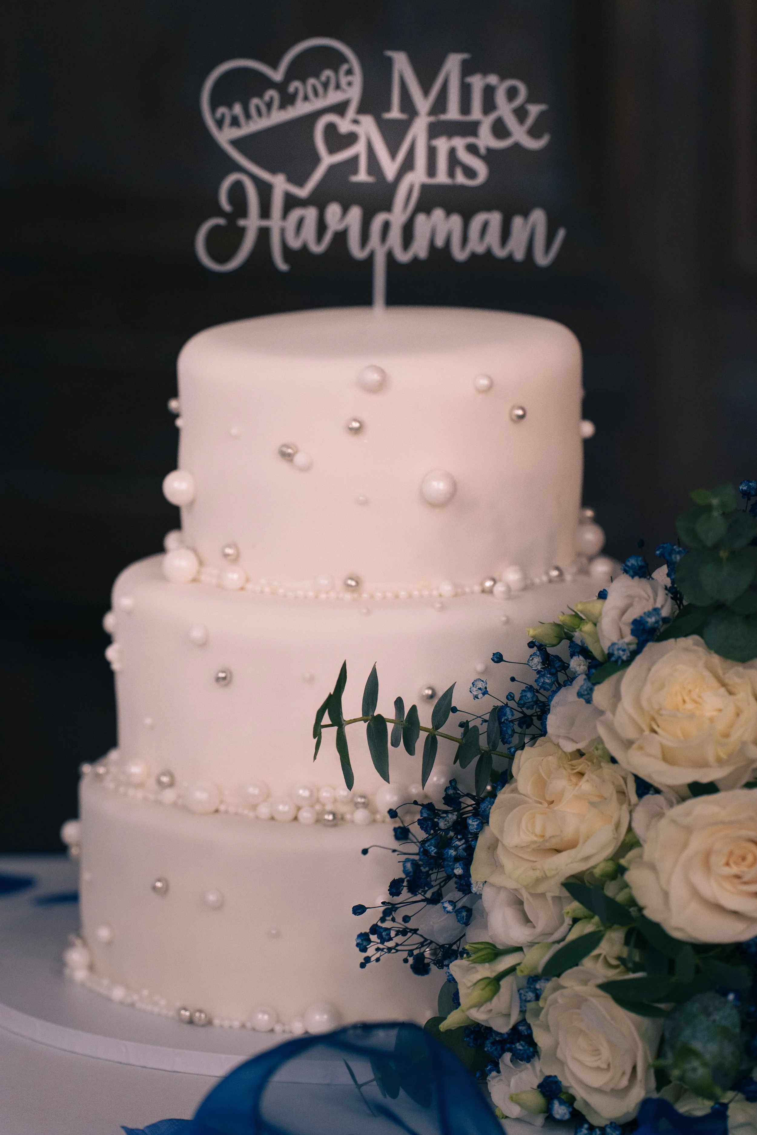 A three-tier white wedding cake decorated with pearl-like beads and a floral arrangement of white roses, blue flowers, and greenery, topped with a laser-cut cake topper that reads "Mr. & Mrs. Hardman" with a heart and date "21.02.2012".