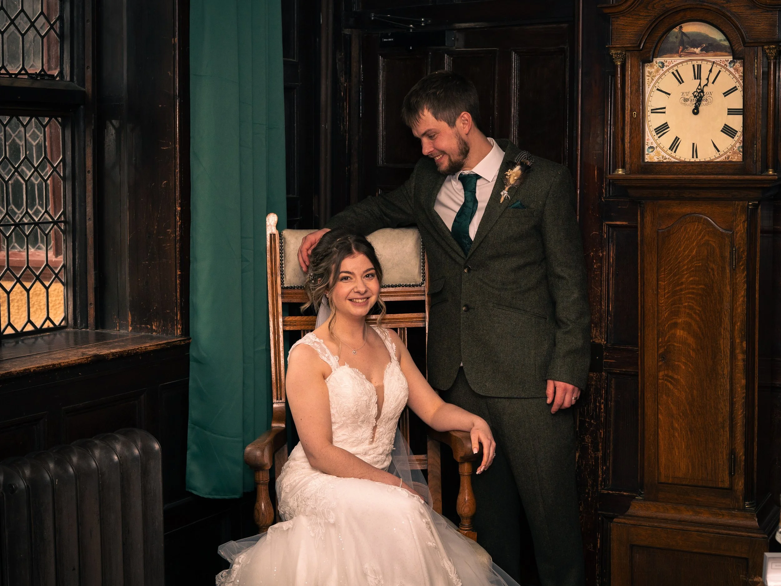 A bride and groom in wedding attire in a vintage-style room with dark wood paneling. The bride is seated in a wooden armchair, wearing a white lace wedding gown. The groom, standing beside her with one hand on her head, wears a dark suit with a shirt