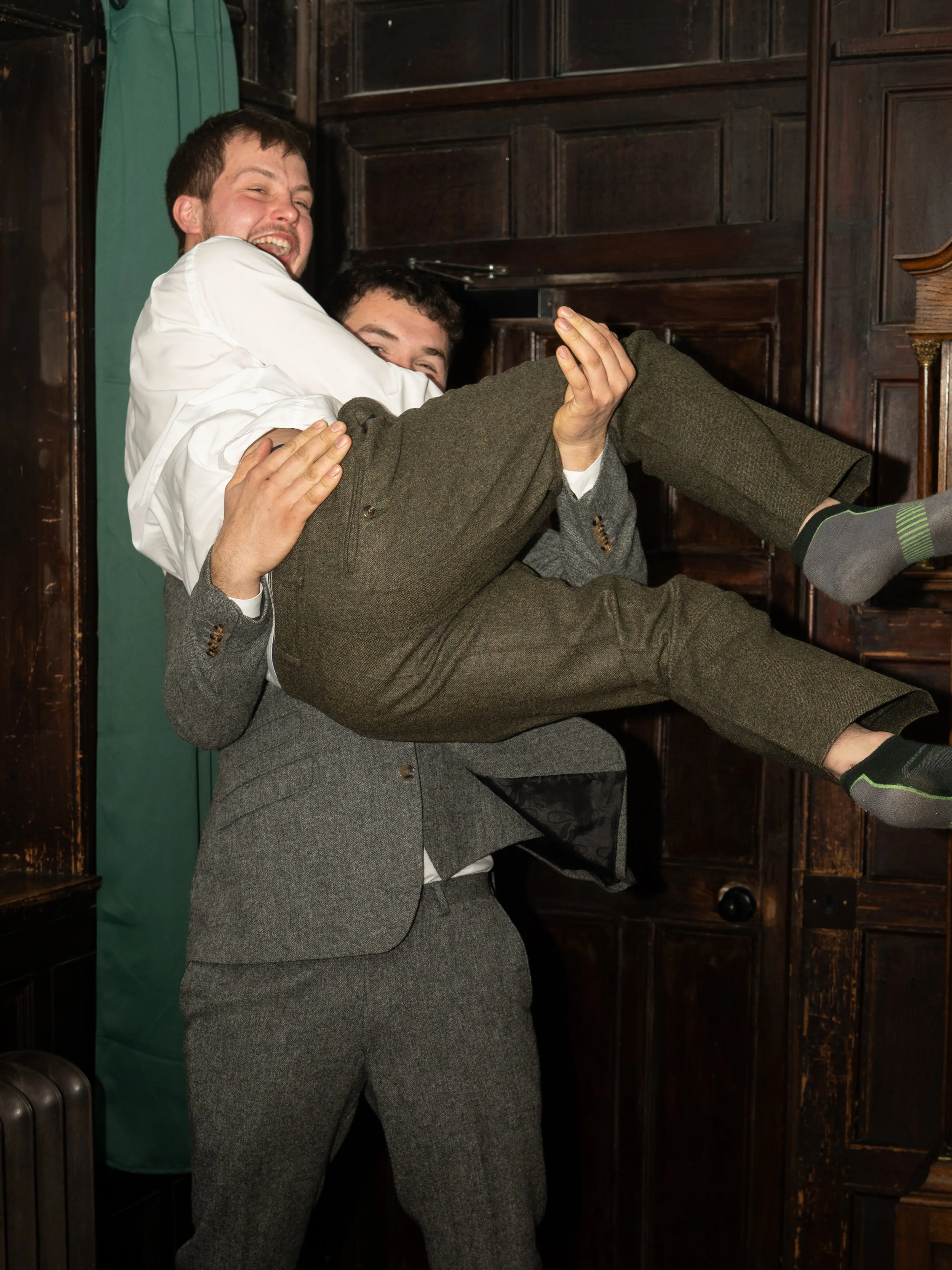 Two men, dressed in formal suits, are having fun and smiling. The man in the tailcoat is holding the other man and lifting him off the ground inside a dark wooden room, possibly a pub or historic building.