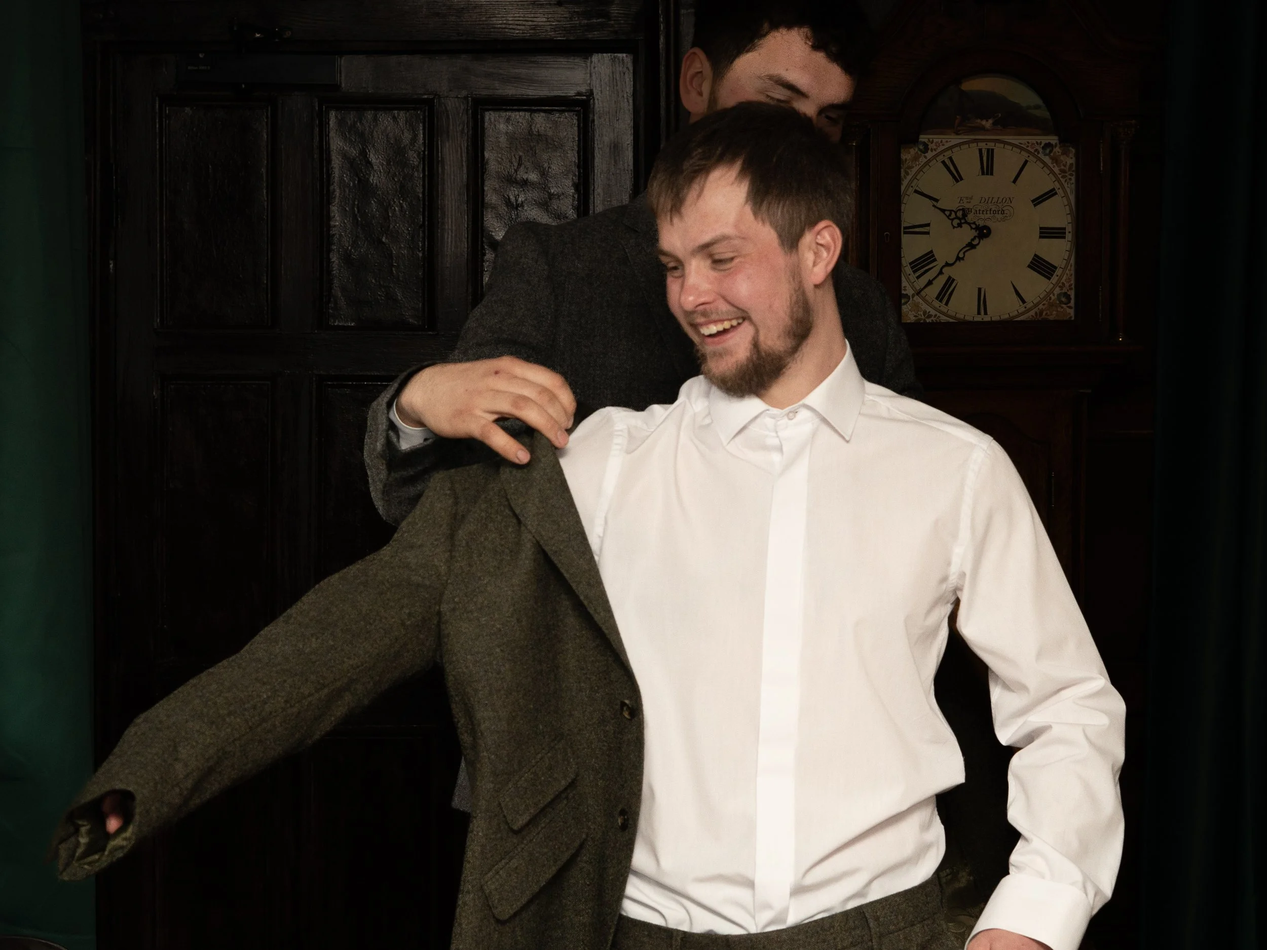 Two men, one in a white shirt, smiling, and the other partially behind him, wearing a blazer. They seem to be in a cozy indoor setting with a vintage clock in the background.
