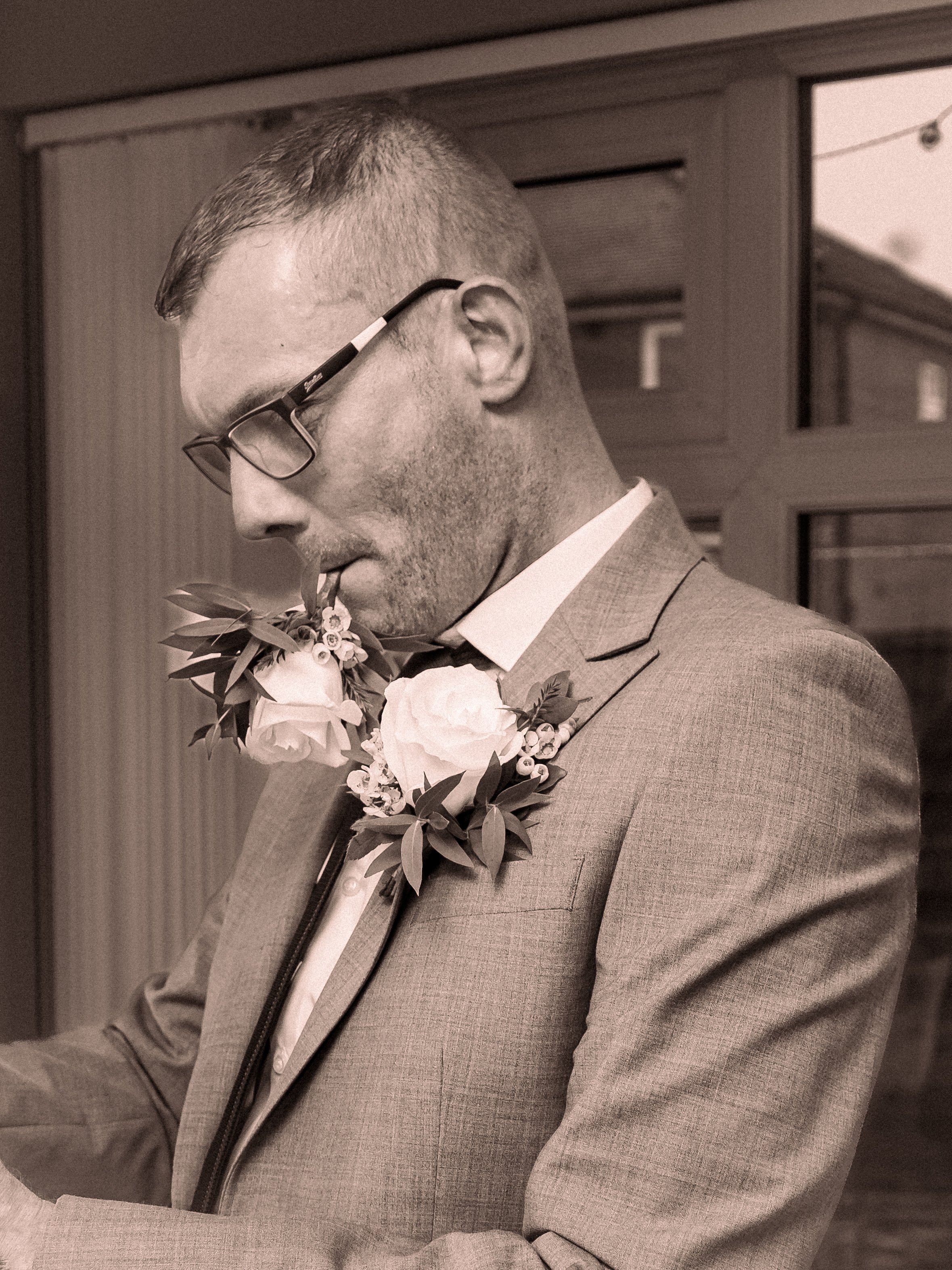 A man in a suit with glasses and a boutonniere, holding a flower, looking down.
