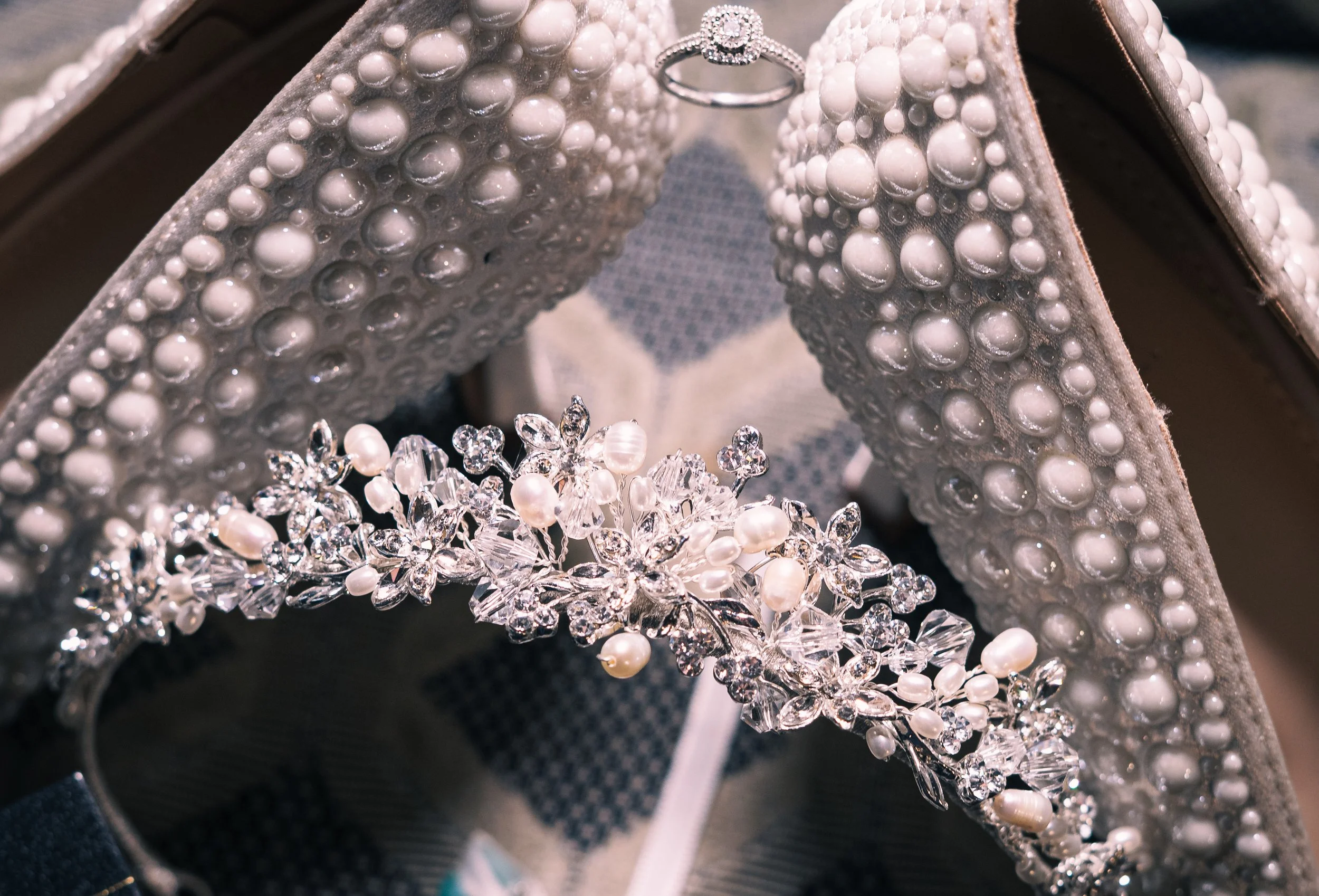 Close-up of bridal accessories including a pearl and crystal headband, a pearl and diamond ring, decorated wedding shoes with pearl and crystal embellishments, all arranged inside an open shoe box.