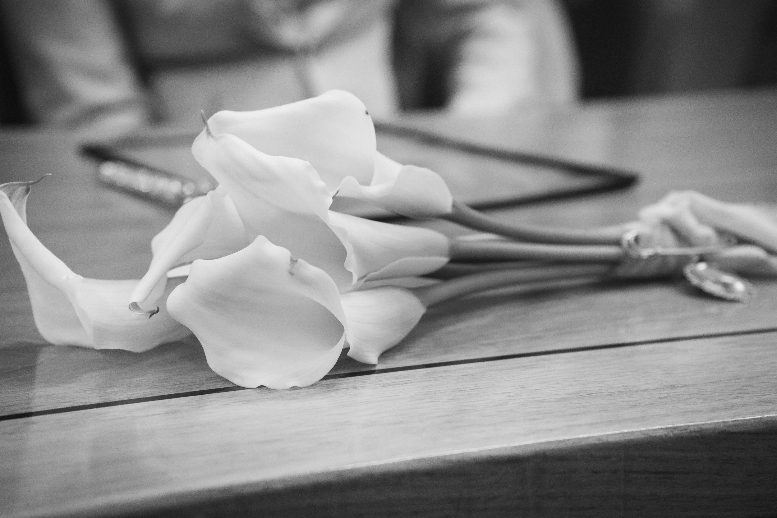 Black and white photo of a bouquet of calla lilies on a wooden table.