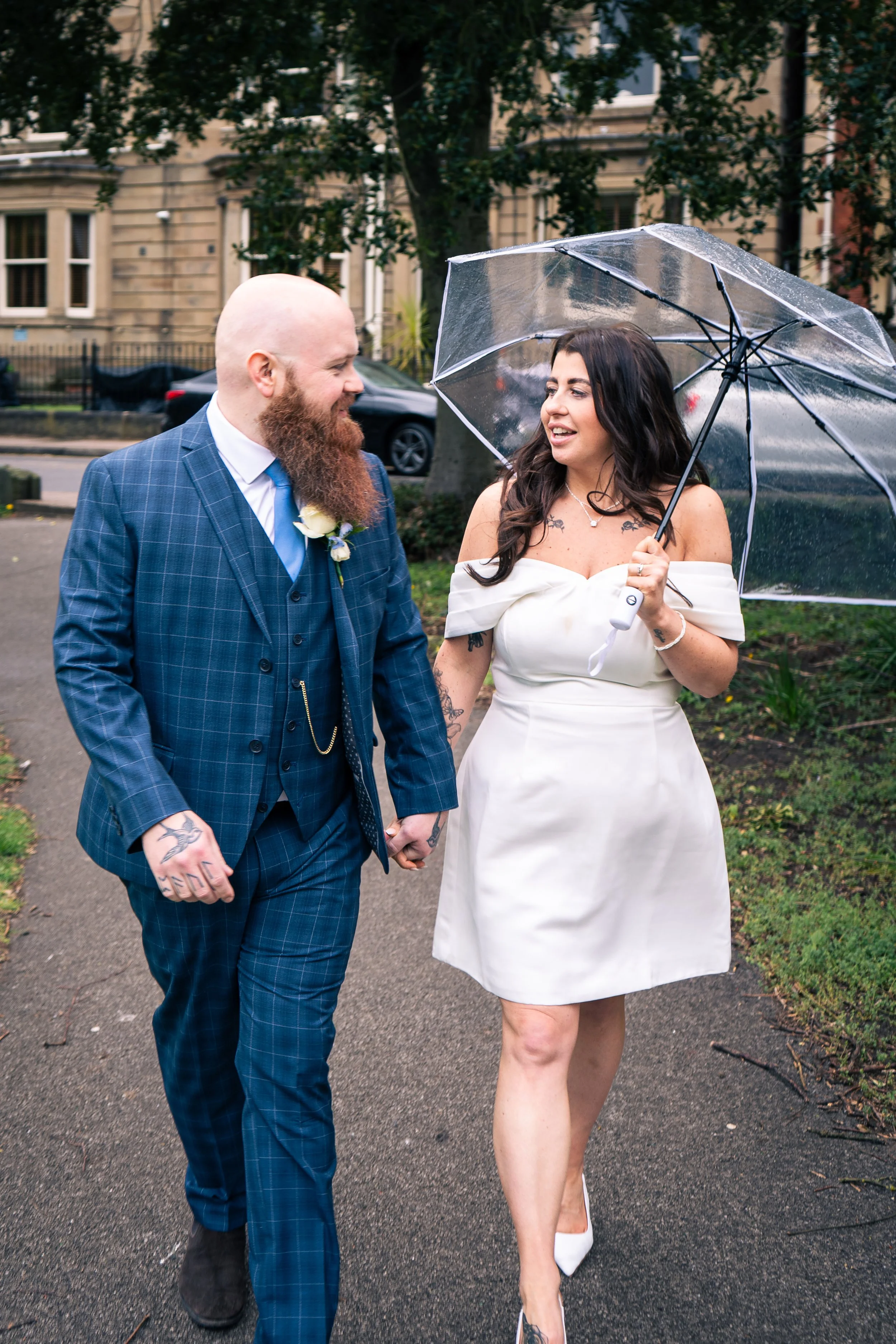 A couple holding hands outdoors on a rainy day, the woman is holding a transparent umbrella, both dressed in wedding attire, smiling at each other.