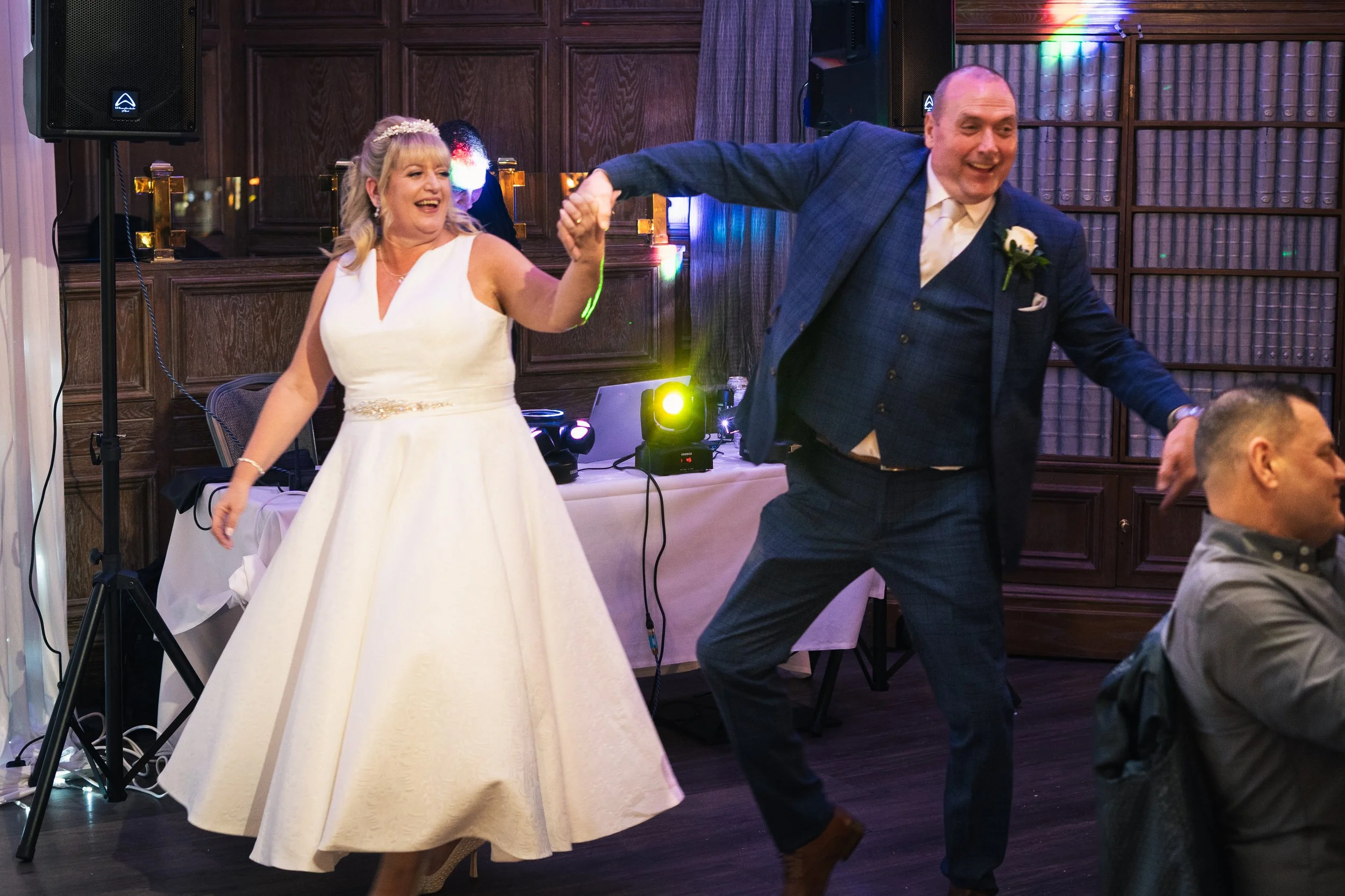 A bride and groom are dancing and holding hands at a wedding reception. The bride is wearing a white wedding dress and the groom is dressed in a blue suit. They are smiling and enjoying the celebration.