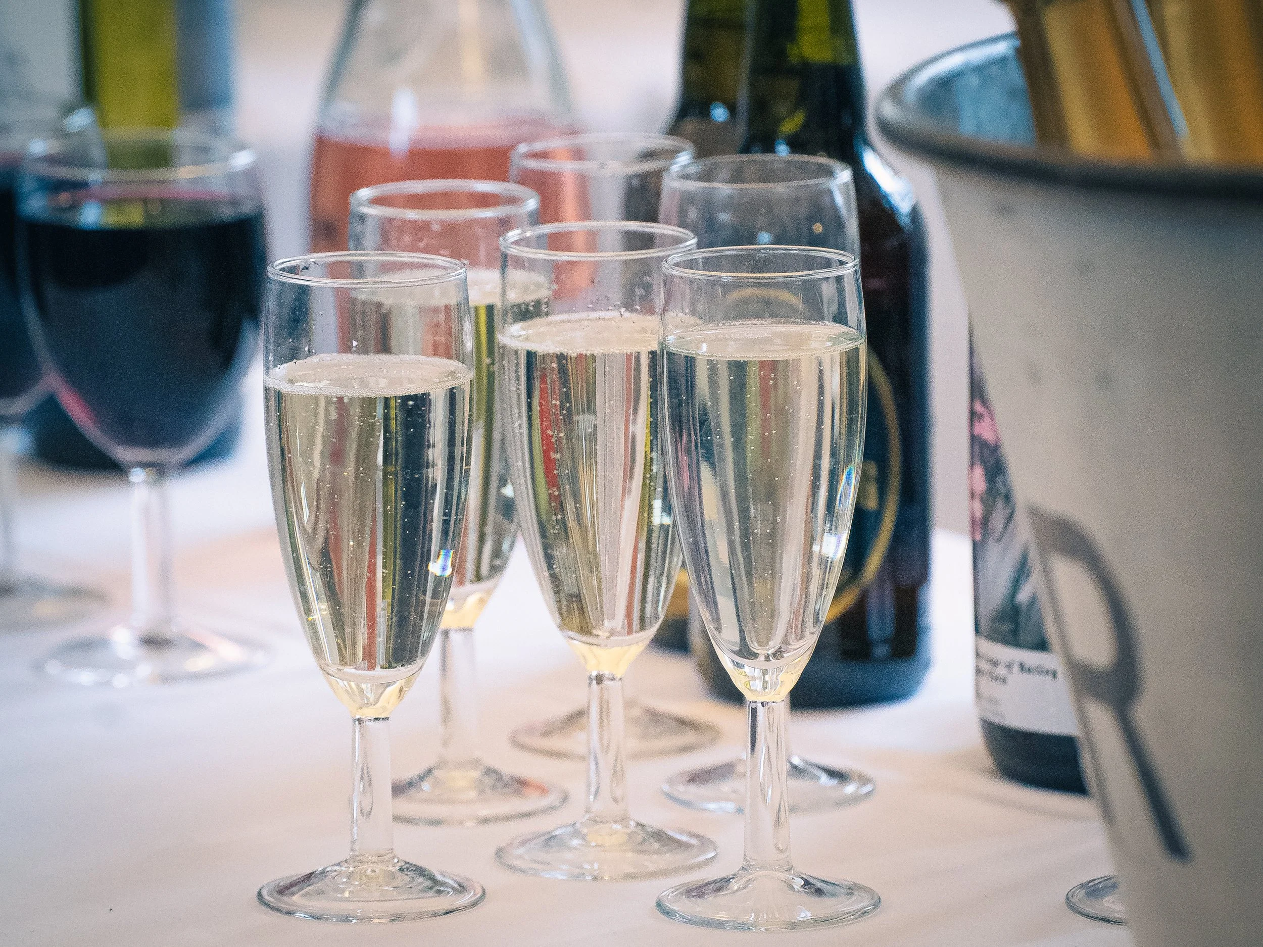 Multiple glasses of champagne and other beverages on a table, with bottles and a bucket in the background.