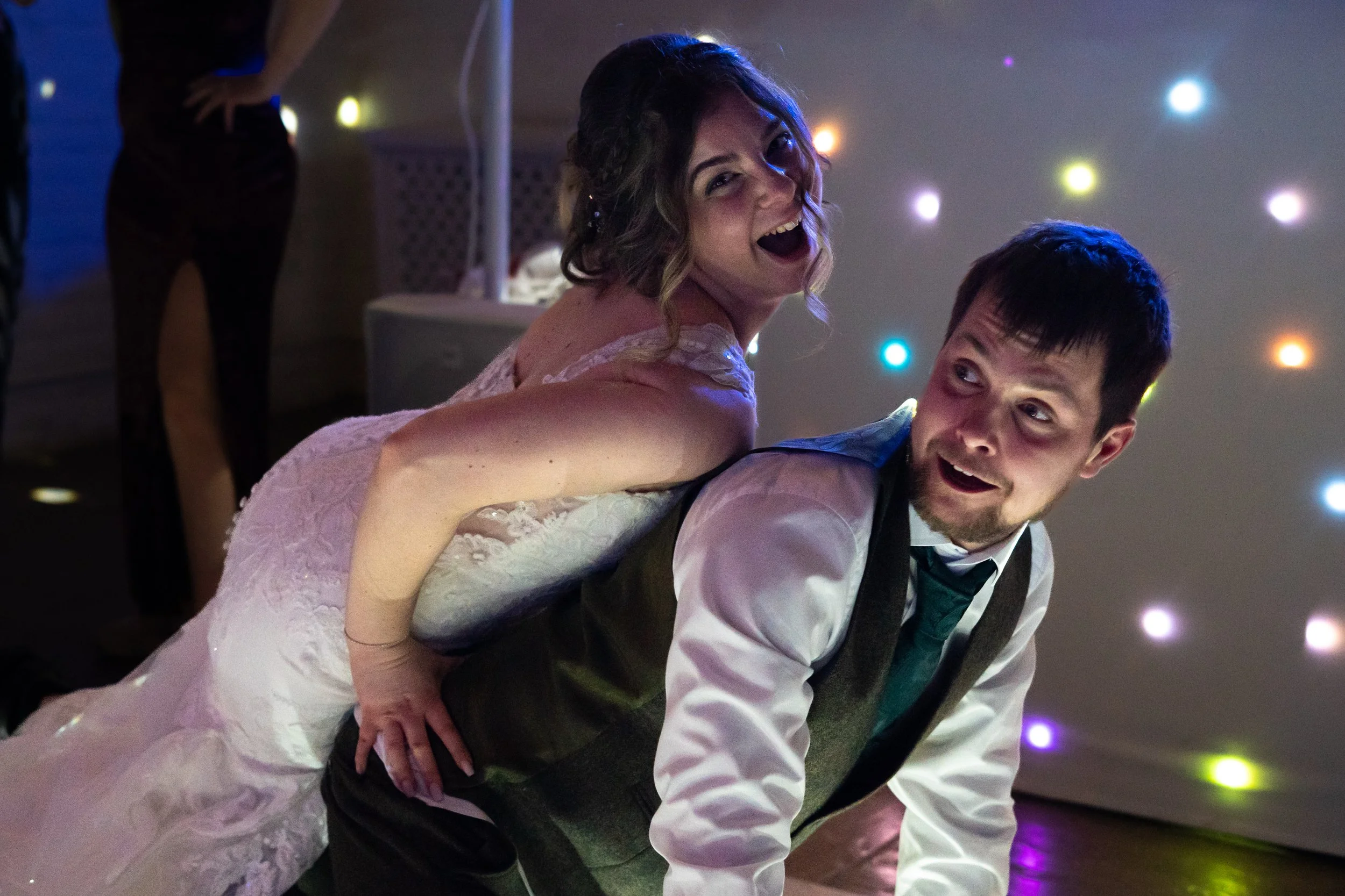 A bride and groom dance at their wedding reception, with the bride on the groom's back, smiling and laughing, surrounded by colorful party lights.