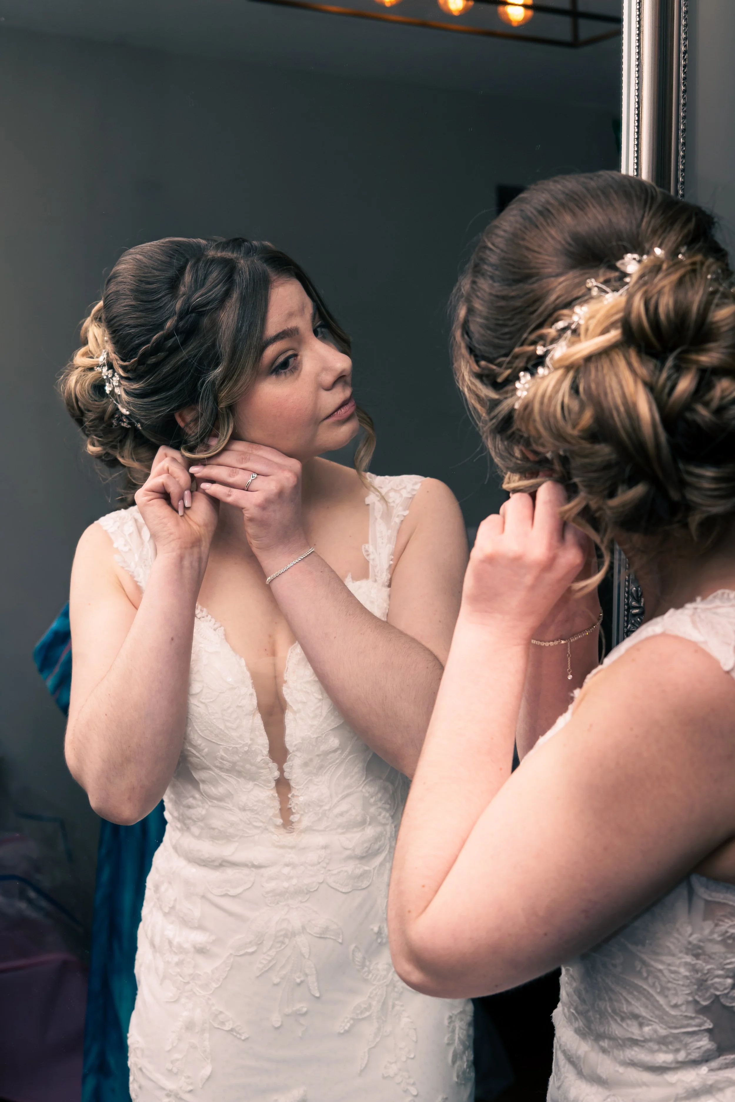 A bride in a lace wedding dress is getting ready, putting on earrings while looking into a mirror.