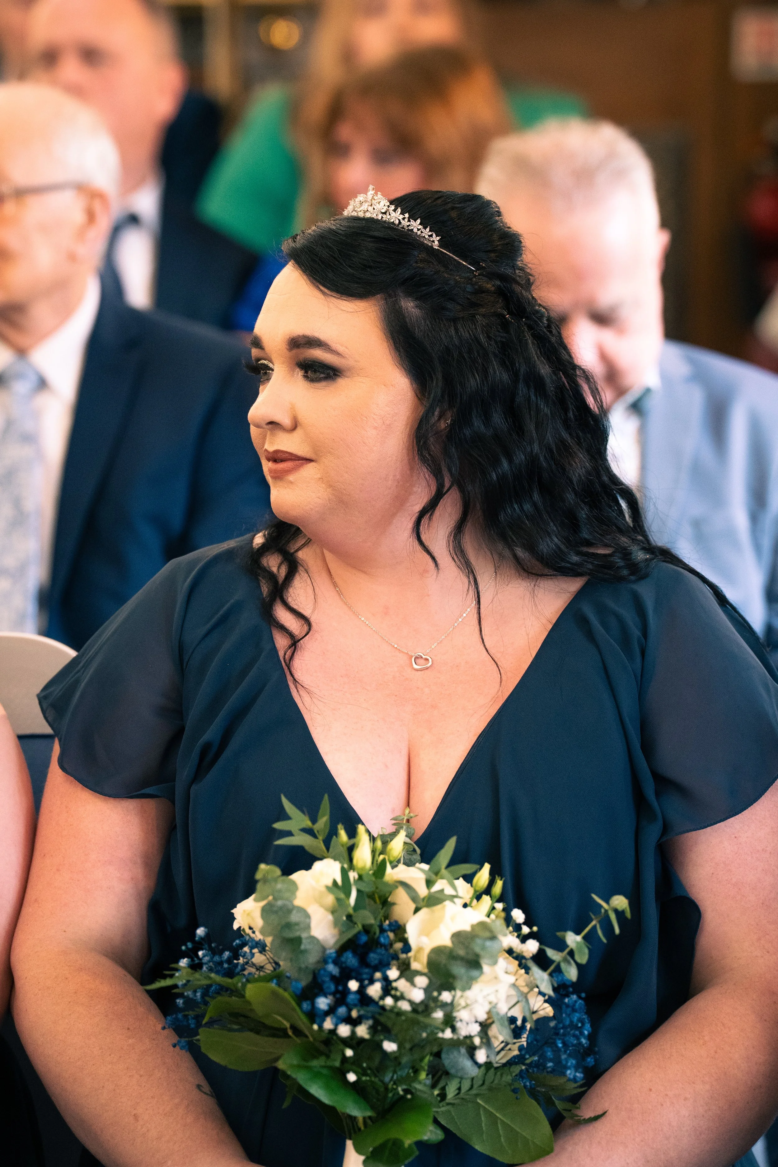 Woman with dark hair wearing a blue dress and tiara, holding a bouquet with white and blue flowers, at a ceremony surrounded by seated people.