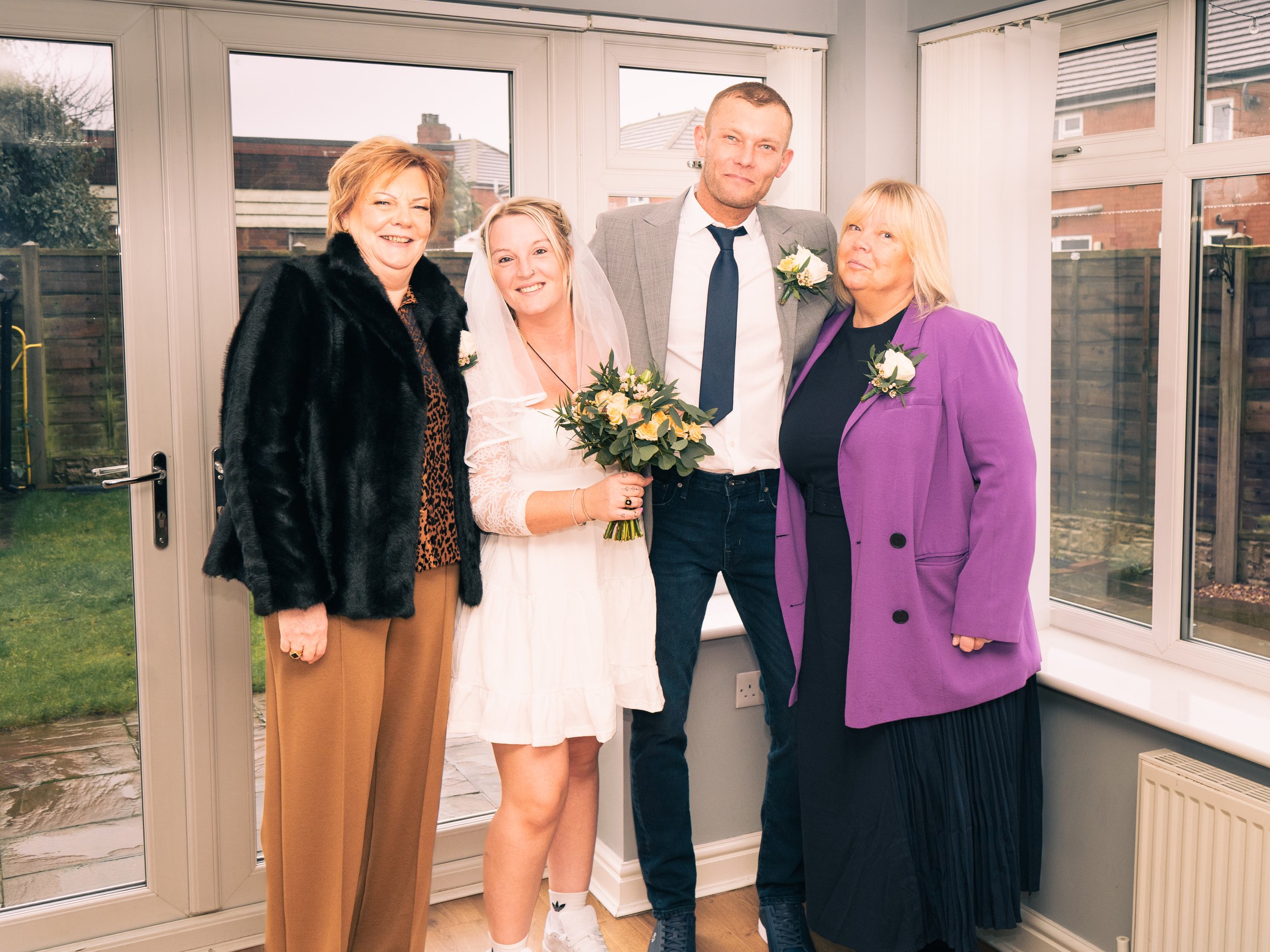 A group of five people at a wedding, including the bride holding a bouquet, two women, and two men, standing inside a house with large windows and a backyard view.