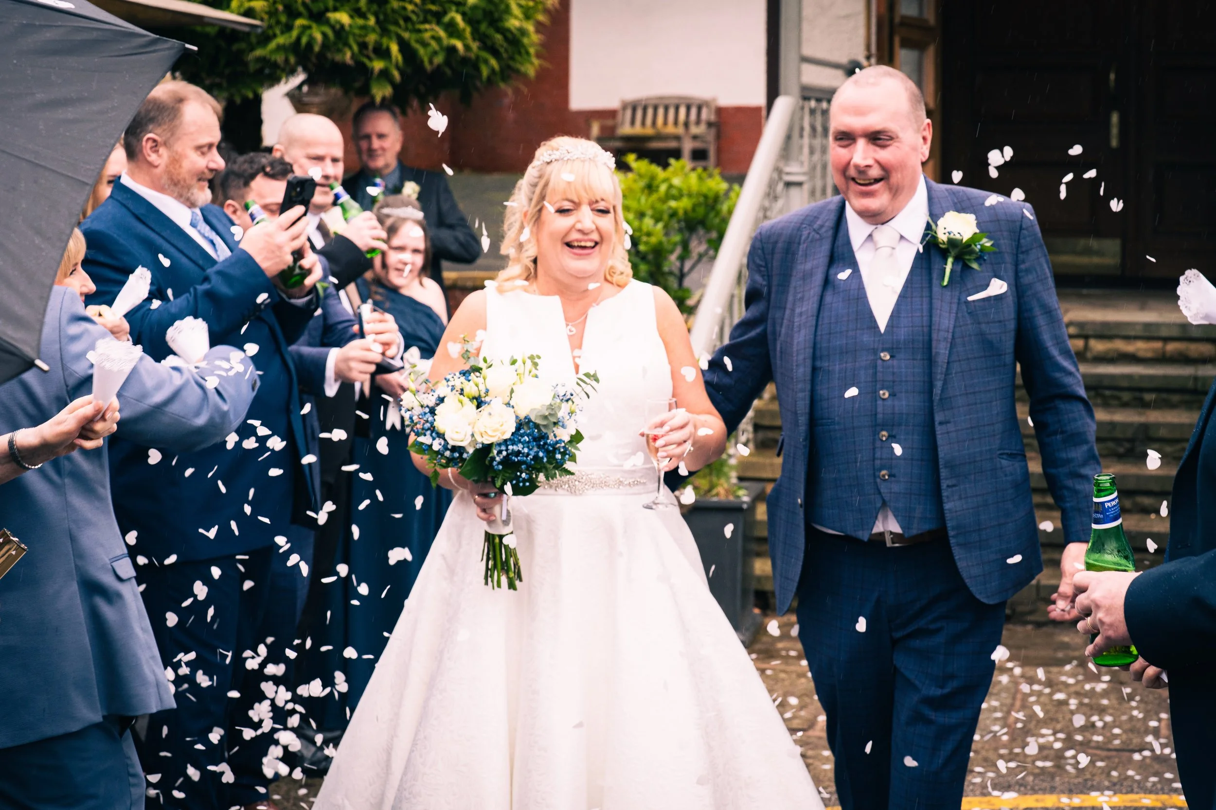 A bride and groom walking hand in hand, smiling, surrounded by wedding guests throwing confetti, outside a house with stairs and greenery.
