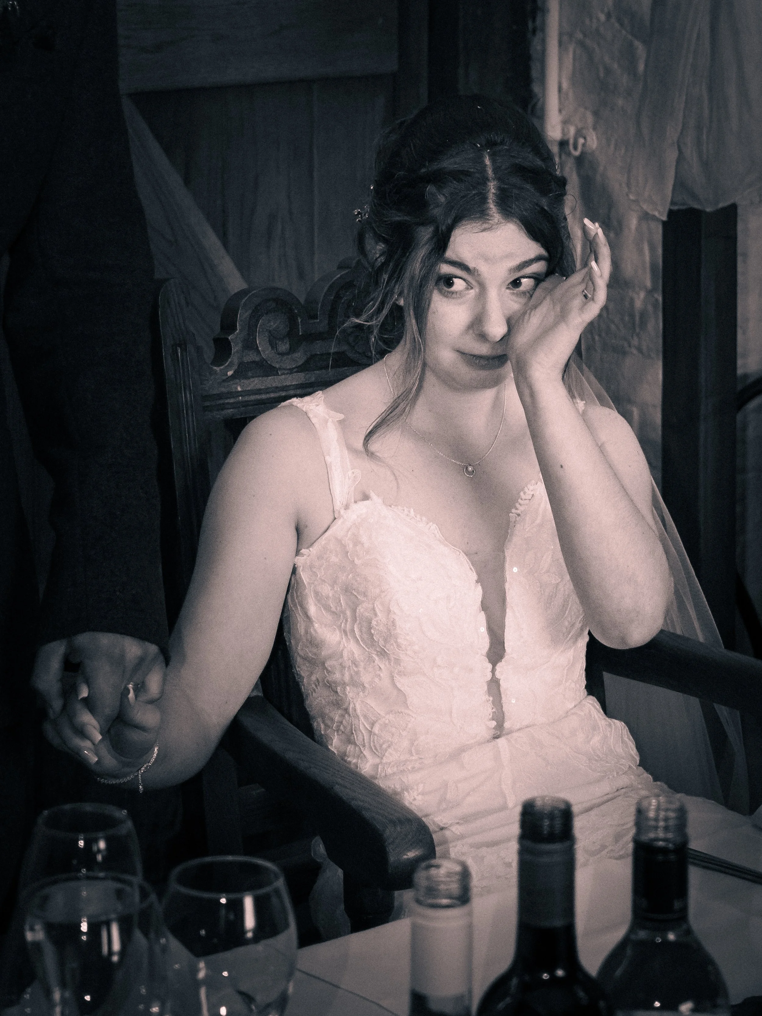 A woman in a wedding dress sitting in a wooden chair at a table, wiping her eye with her hand, with bottles and glasses on the table.
