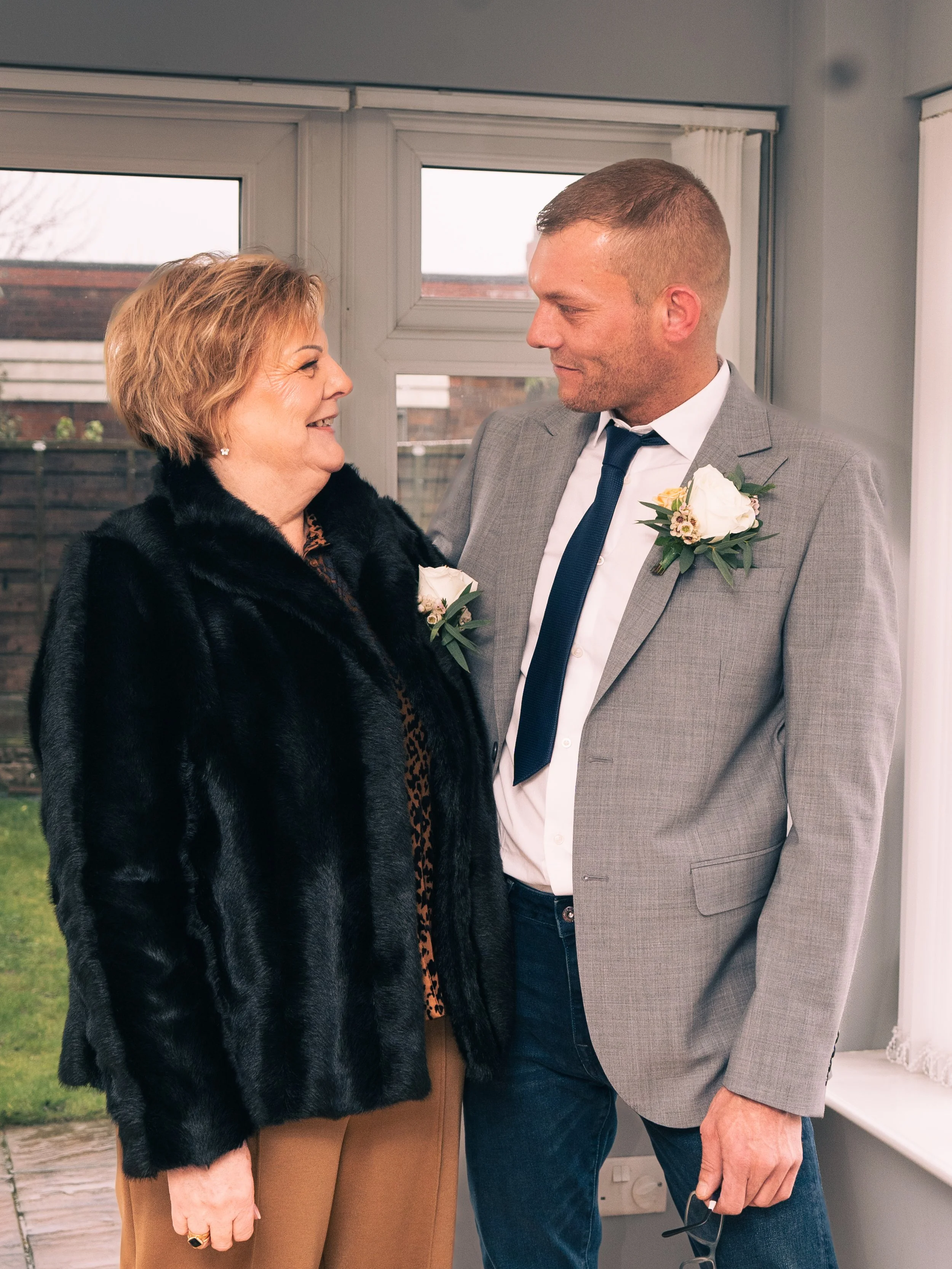An older woman and a young man are smiling at each other, inside a room with a window behind them. The woman is wearing a black fur coat, brown pants, and has short blonde hair. The man is wearing a gray suit with a white dress shirt, a dark tie, and