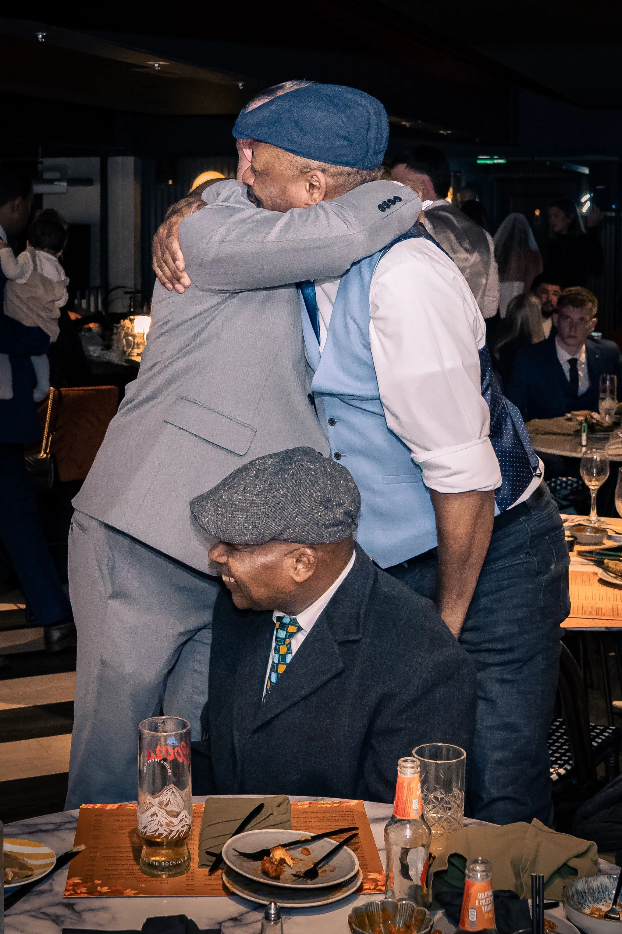 Two men embracing at a social gathering, one wearing a light gray blazer and the other wearing a blue vest and white shirt, while a man in a gray cap and dark blazer sits in the foreground.
