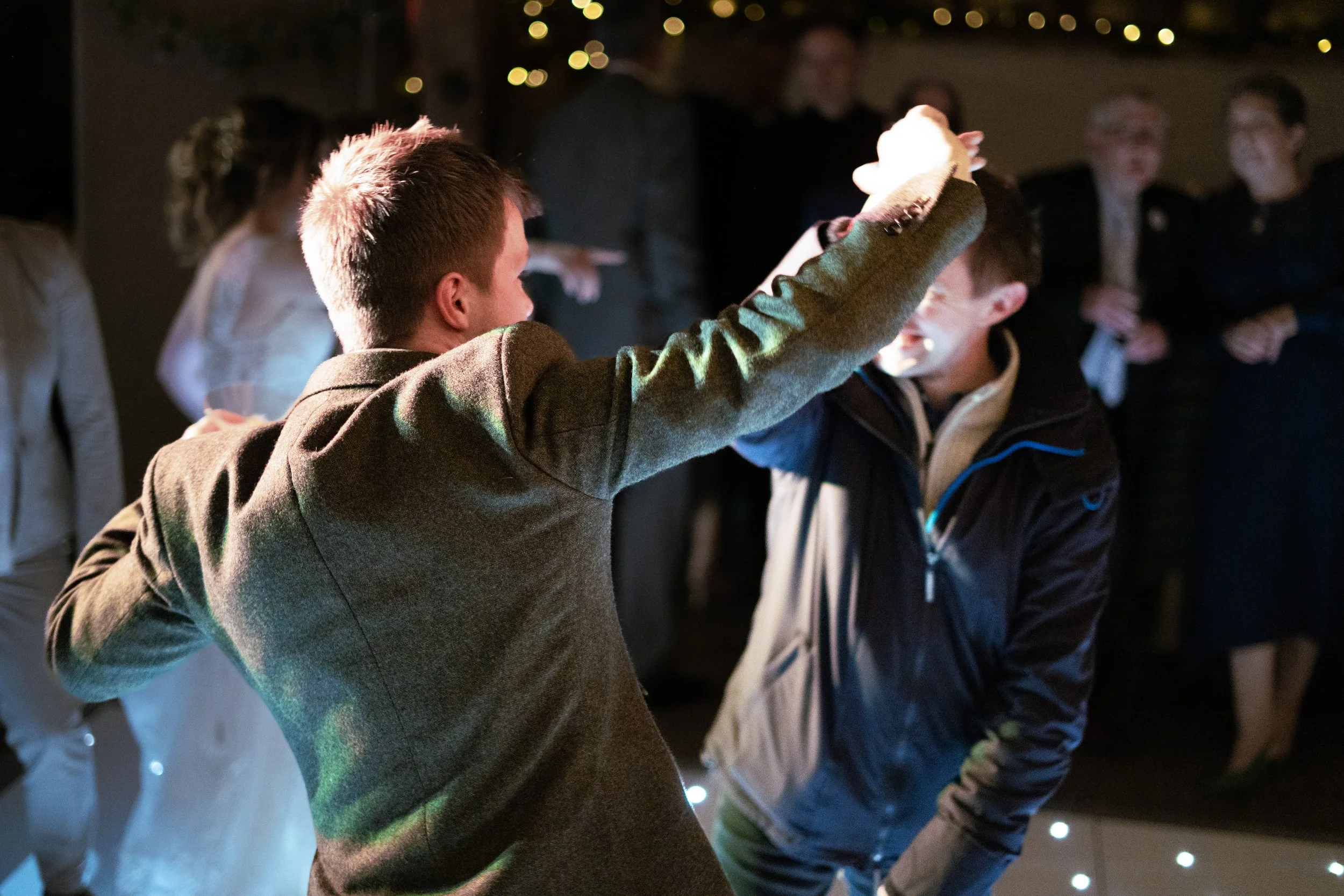 Two boys are dancing and smiling at a party, surrounded by adults in the background. One boy is wearing a brown blazer, and the other is wearing a black jacket. The scene is festive with dim lighting and string lights in the background.