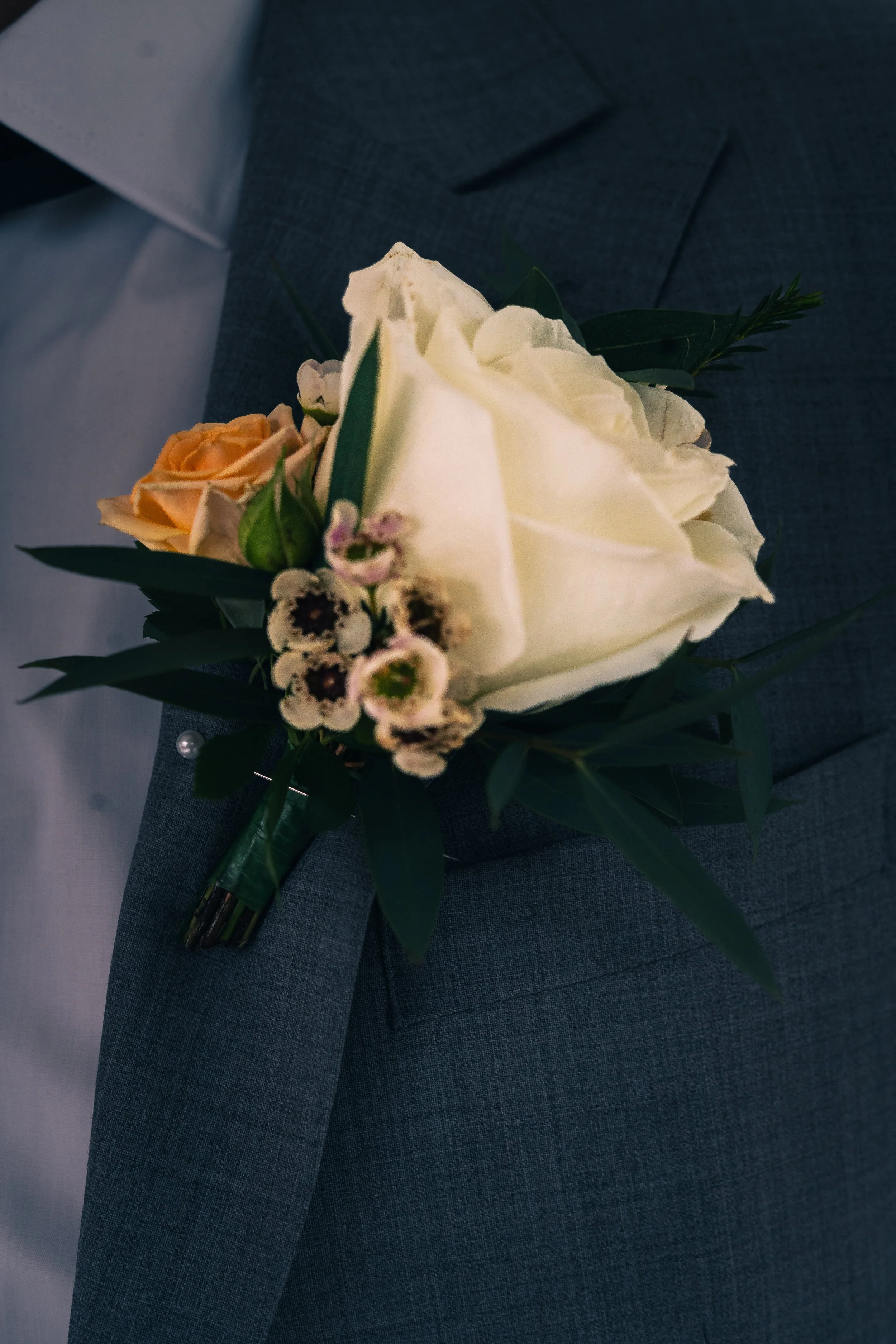 A boutonniere with a white rose, small peach rose, and tiny white flowers pinned to a gray suit jacket.