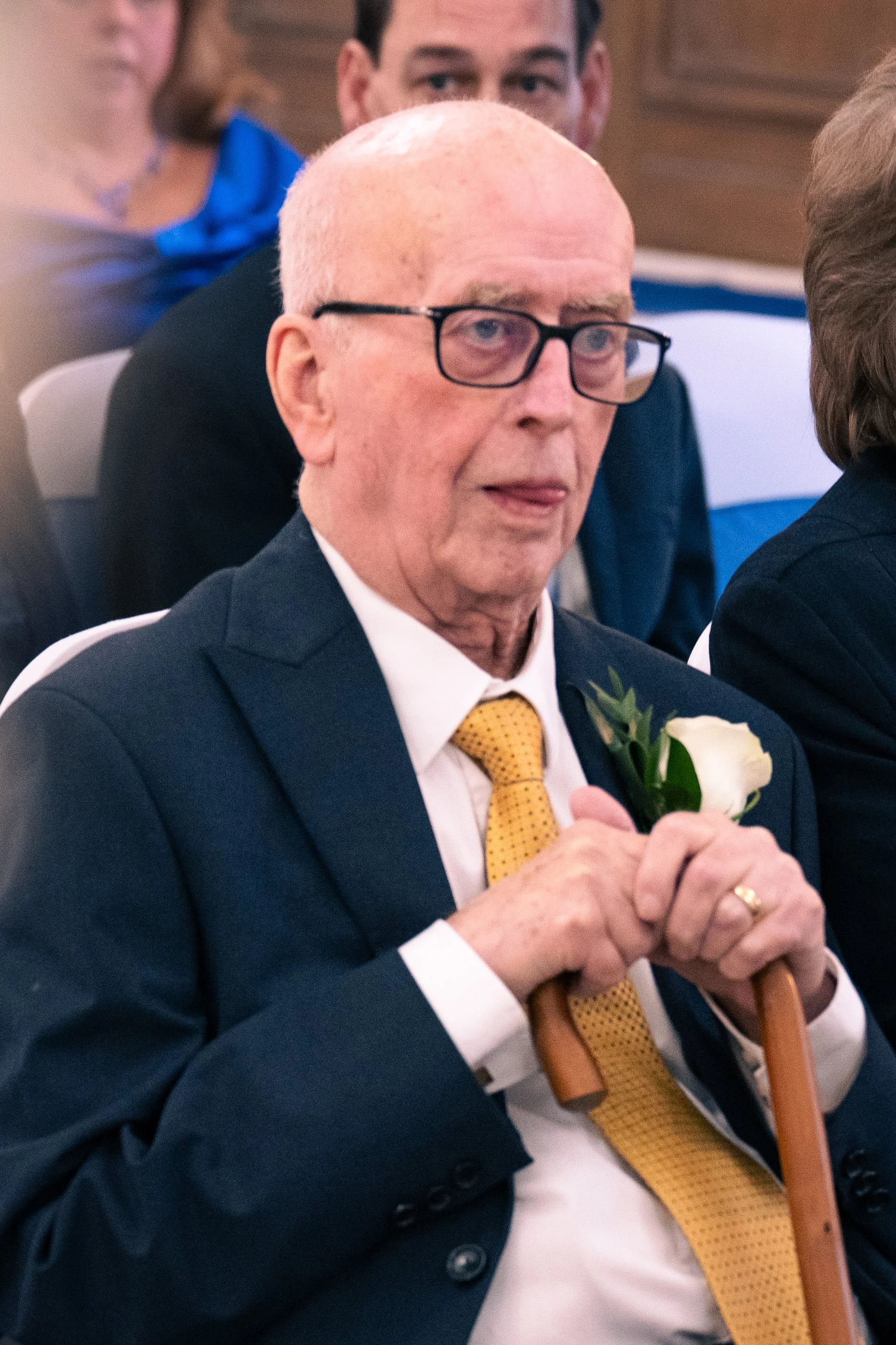 An elderly man with glasses, dressed in a dark suit, white shirt, and a yellow tie, sitting at an indoor event, holding a cane and a white flower pin on his lapel.