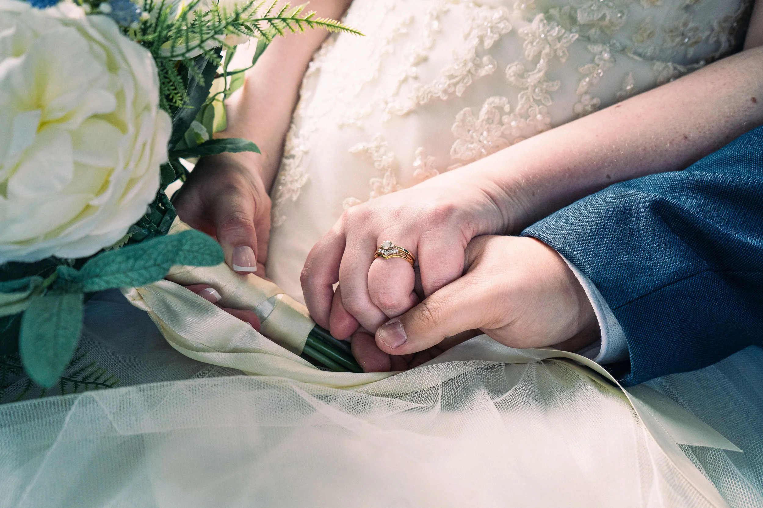 Close-up of a bride and groom holding hands during their wedding, featuring wedding rings, bridal dress, groom's suit, and bouquet.