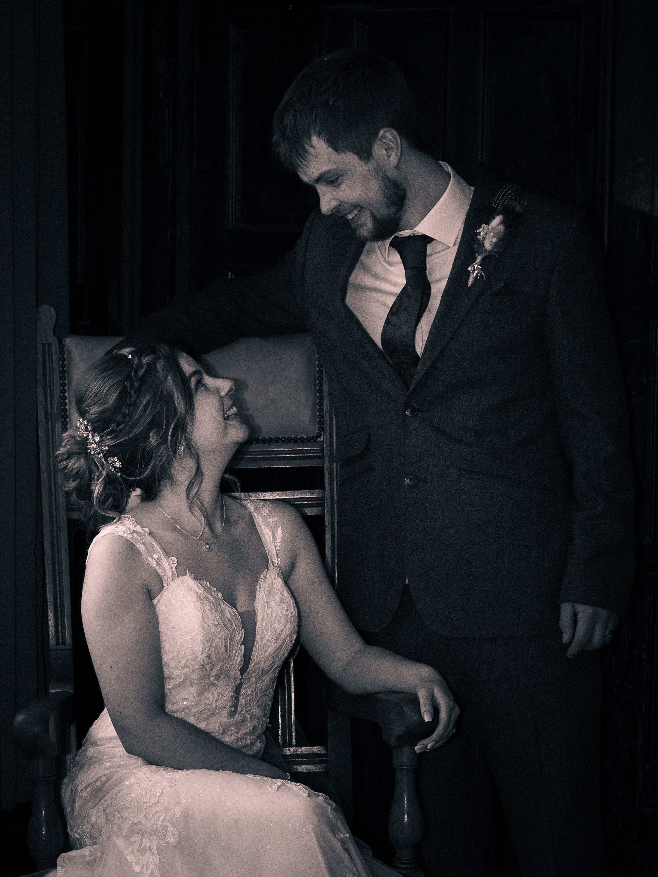 A man in a suit leaning over a seated woman in a lace dress, both smiling at each other affectionately in a dimly lit room.