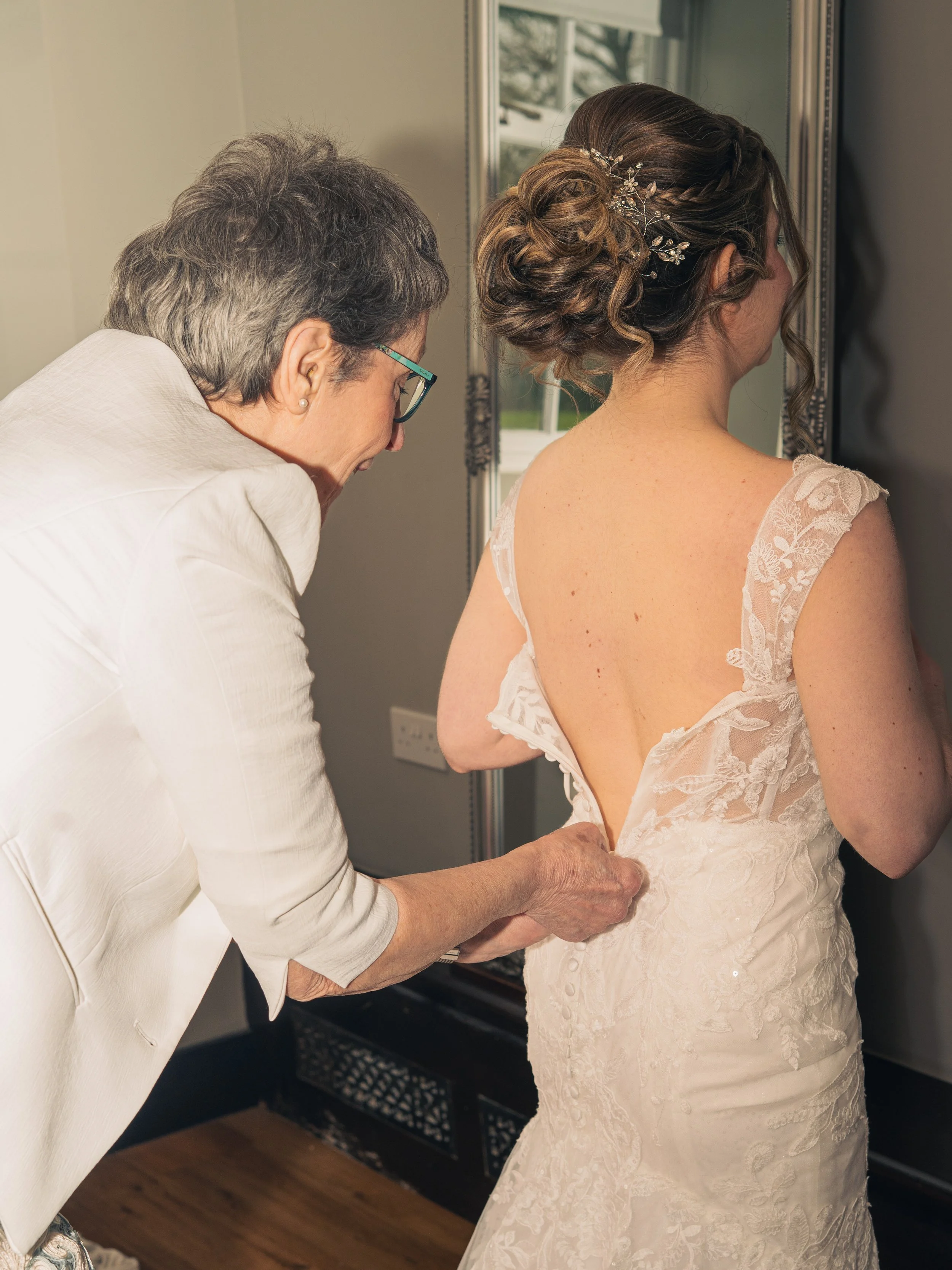 A woman is helping a bride put on her wedding dress, showing the back details of the gown.