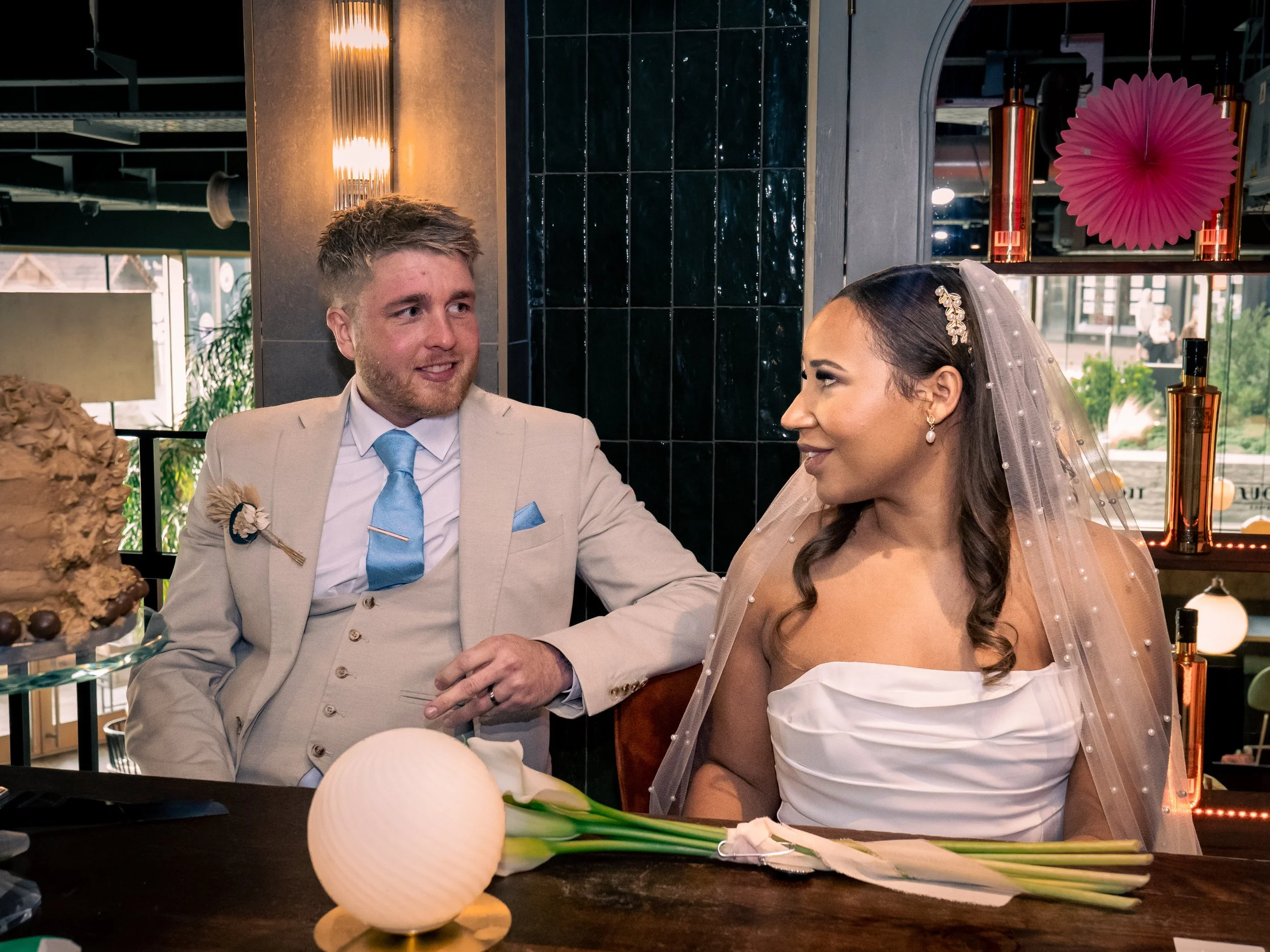 A newly married couple sitting at a table, the groom in a beige suit with a blue tie, and the bride in a strapless white wedding dress with a veil and jewelry, smiling at each other during their wedding celebration.