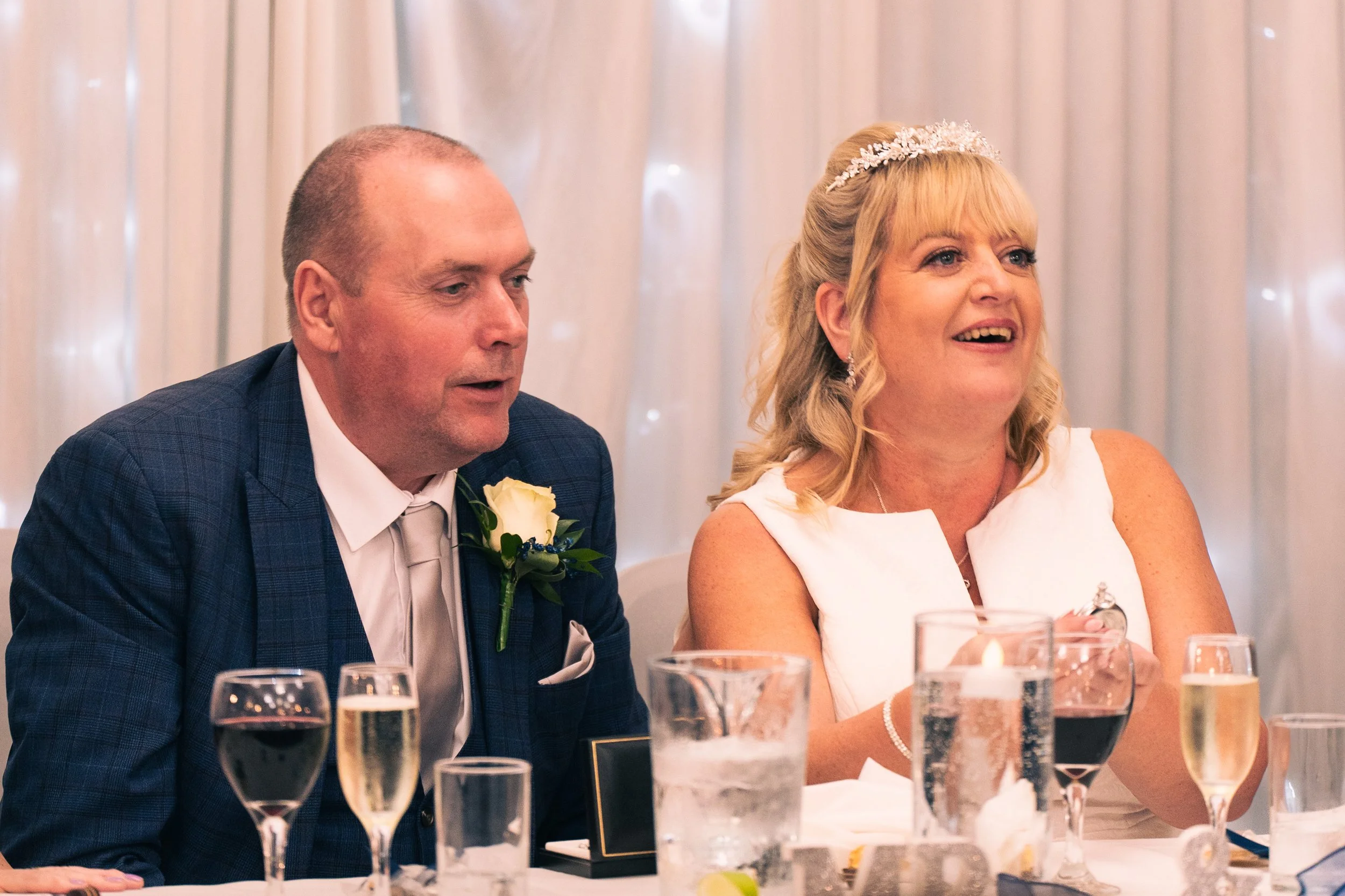 A woman in a white dress with a tiara, sitting at a wedding reception table, smiling and clapping. A man in a blue suit with a white boutonniere sits next to her, looking in the same direction.