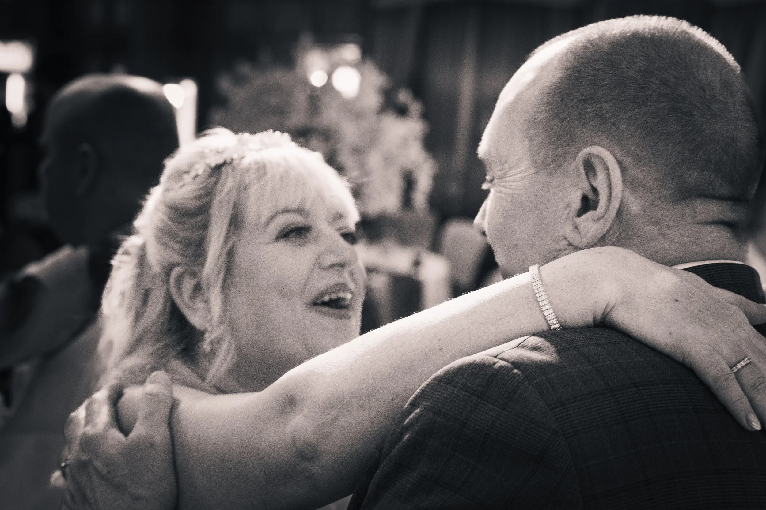A woman with light hair and a bracelet, smiling, embraces a man with glasses and a tuxedo, at what appears to be a happy event or celebration.