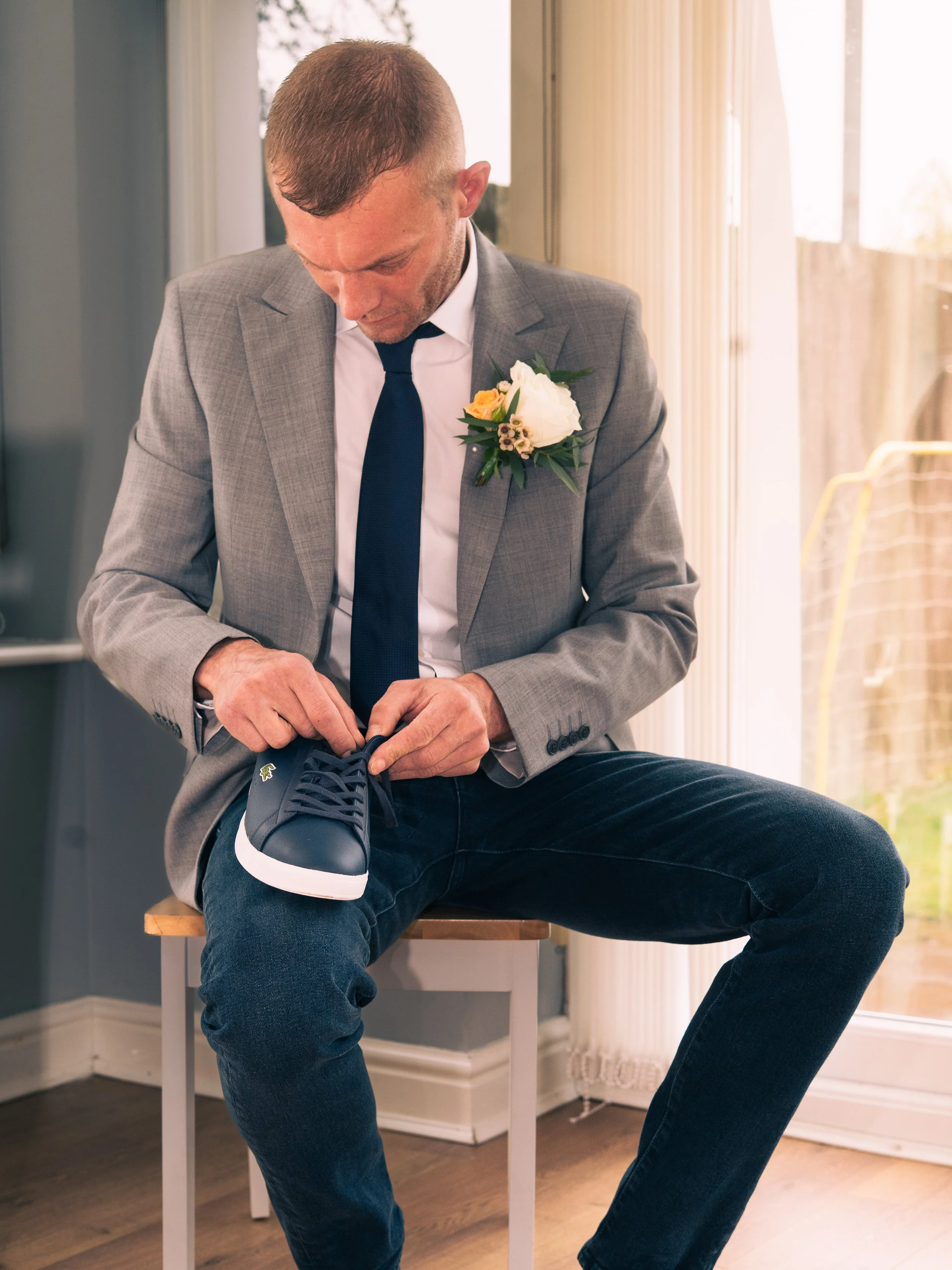 A man in a gray suit jacket, white shirt, and blue tie sitting on a stool, tying his navy blue sneaker. He has a white flower boutonniere on his jacket lapel and is positioned near a window with vertical blinds, in a room with wooden flooring.