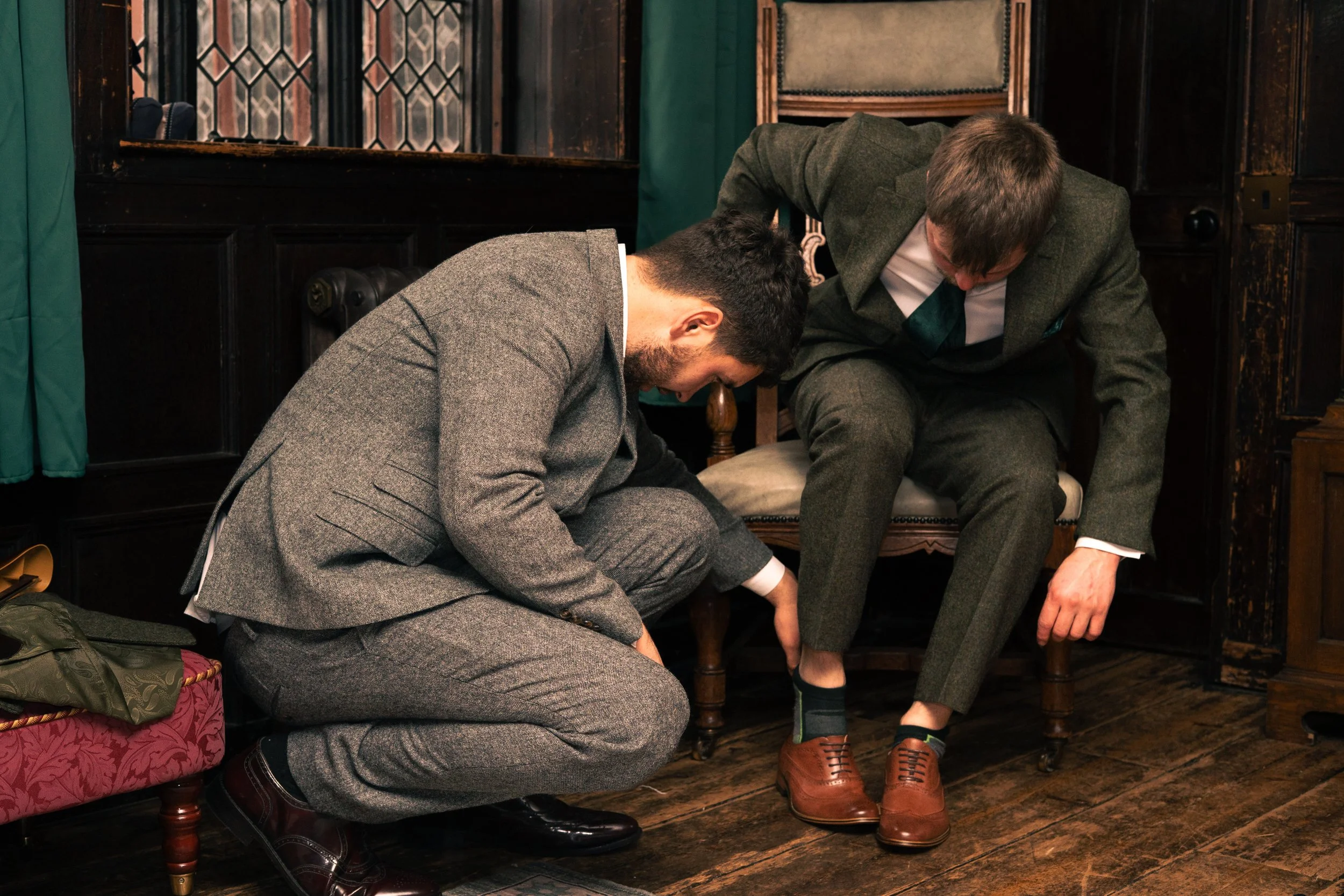 Two men in suits helping each other put on socks in a vintage room with dark wood paneling.