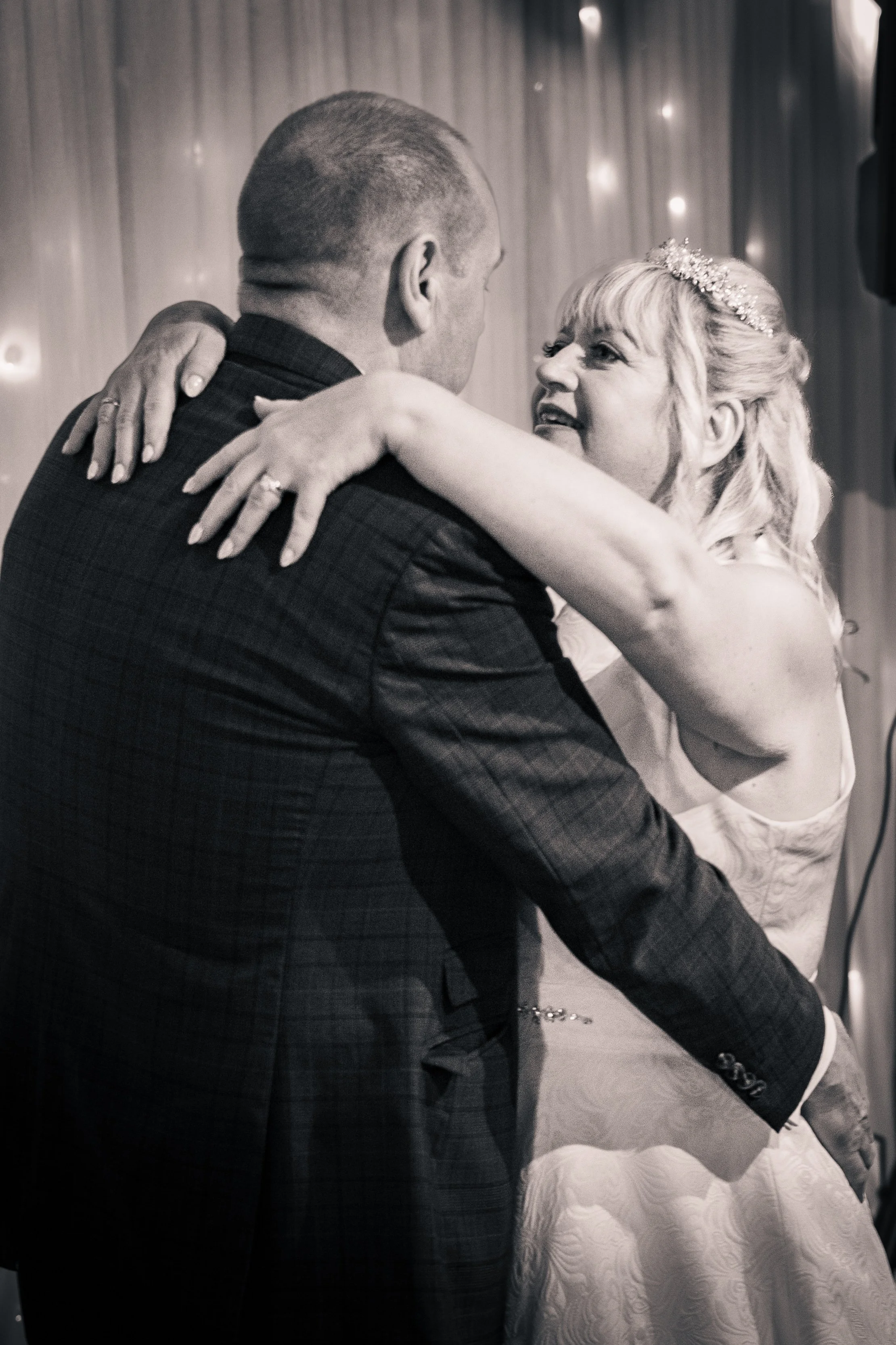 A woman and a man are dancing closely at a wedding reception. The woman has a tiara and a wedding dress, and the man is wearing a dark suit. They are smiling and looking into each other's eyes.