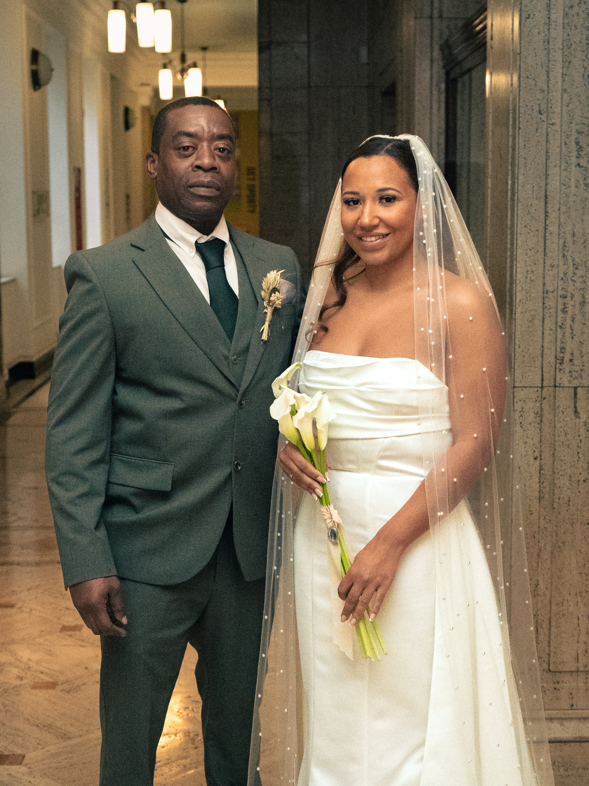 A bride in a white wedding gown holding a bouquet of lilies, standing beside a man in a grey suit, in an indoor setting.