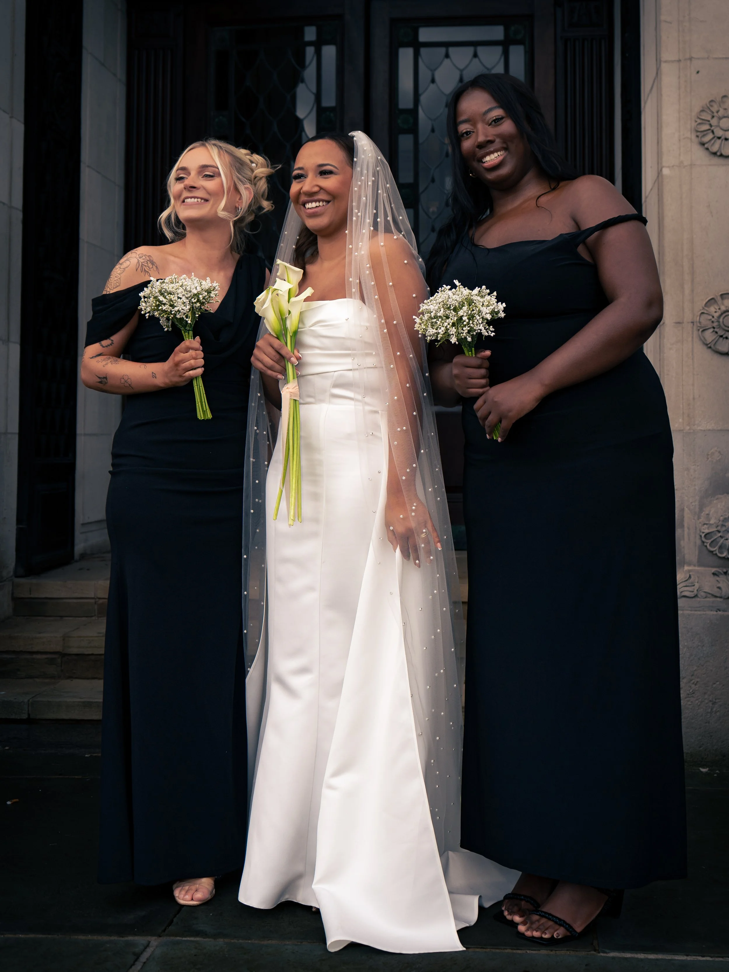 A bride in a white wedding gown and veil stands between two women in black dresses, holding small bouquets of white baby’s breath flowers, outside a building with ornate stonework.