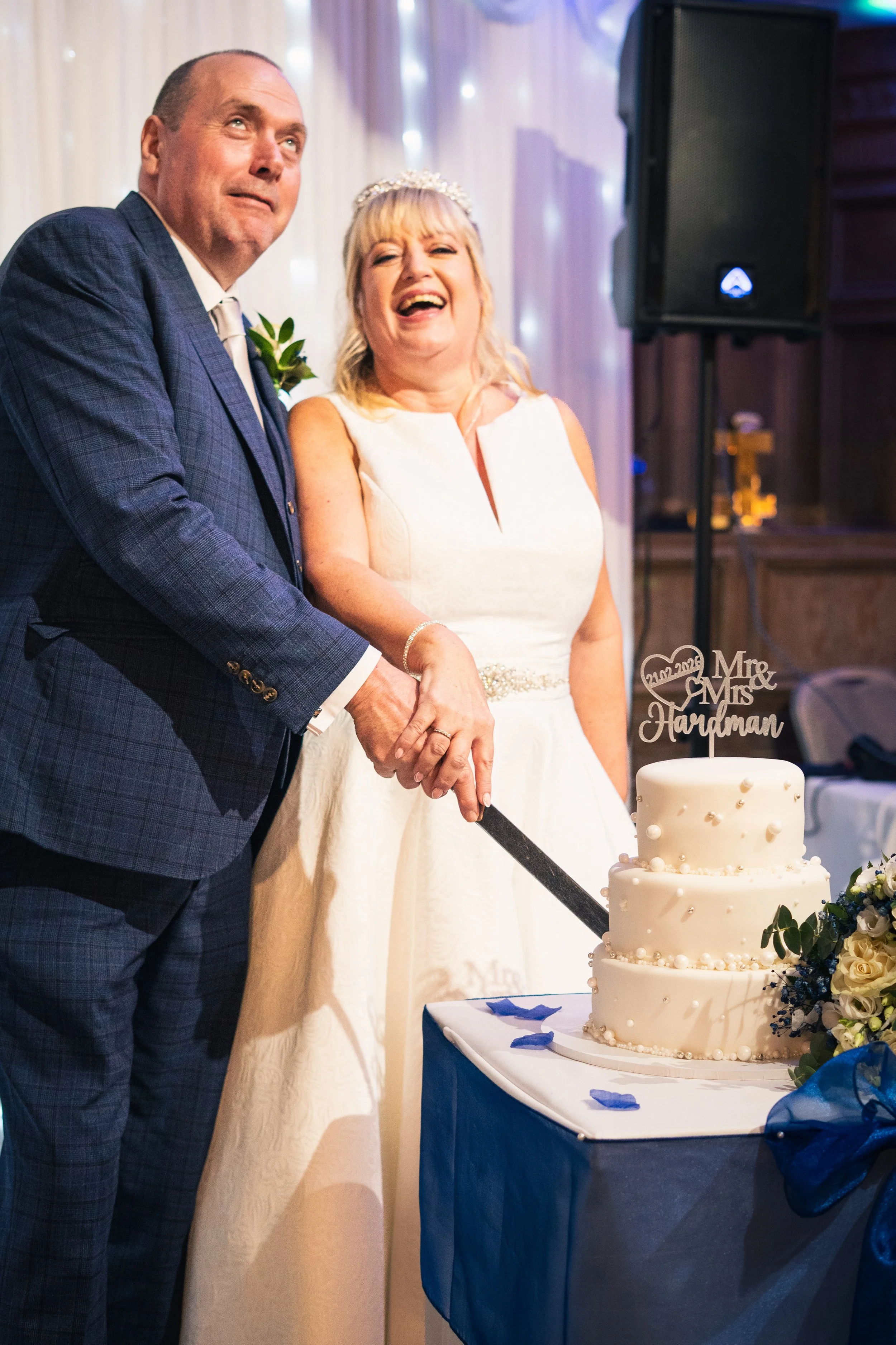 A newlywed couple is cutting a wedding cake together. The bride is laughing and wearing a white dress with a tiara, while the groom is in a suit. The wedding cake has three tiers with white icing and a 'Mr. & Mrs. Handyman' cake topper. The scene is 